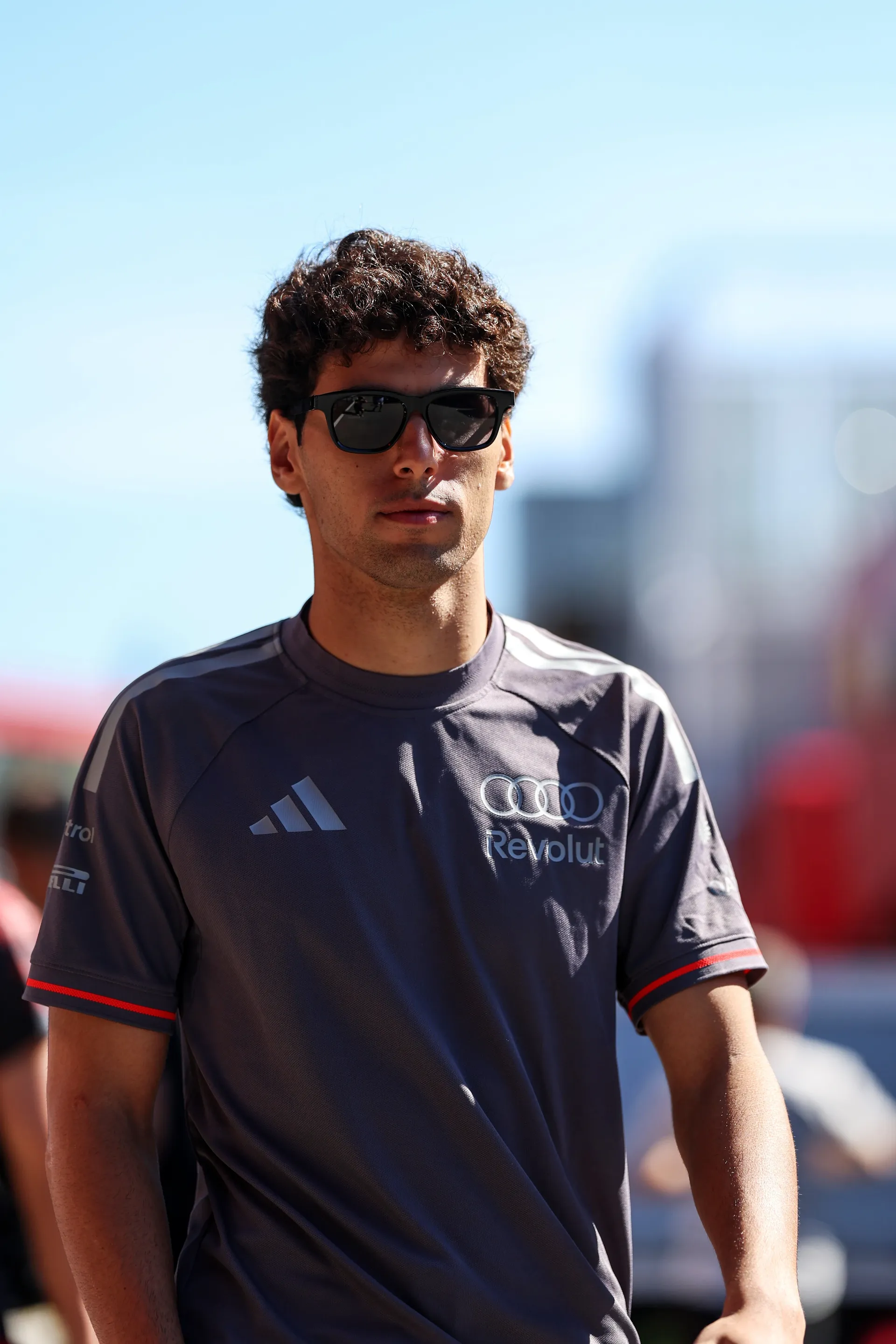 Gabriel Bortoleto walking through the paddock wearing Audi Revolut F1® Team kit and sunglasses, with a blurred circuit background.