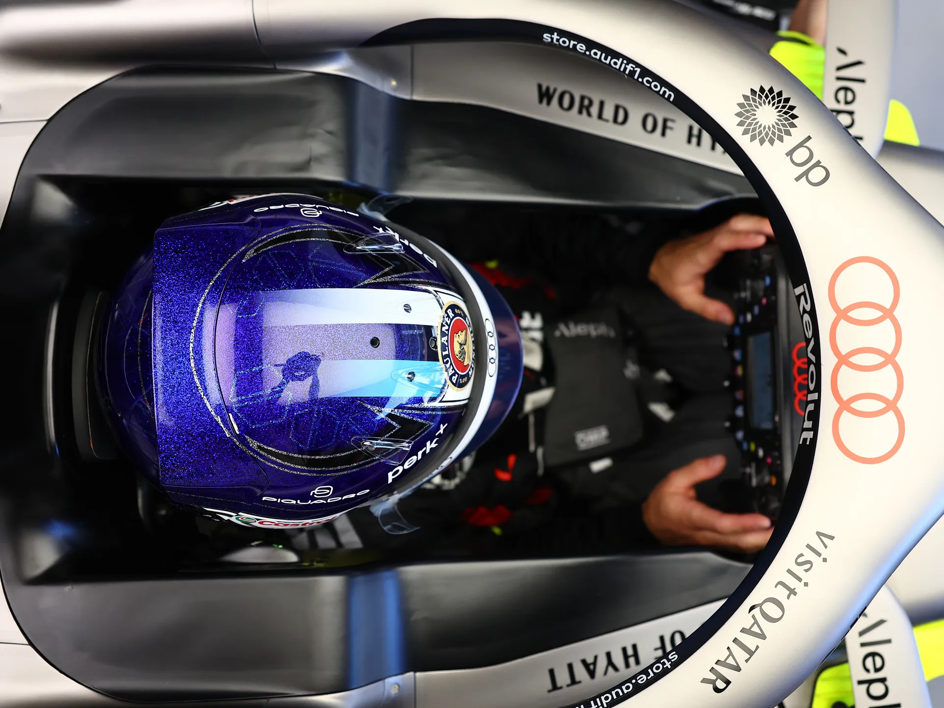 Top-down view of Paul Aron seated in the Audi Revolut F1® Team car cockpit, wearing a blue helmet and hands on the steering wheel.