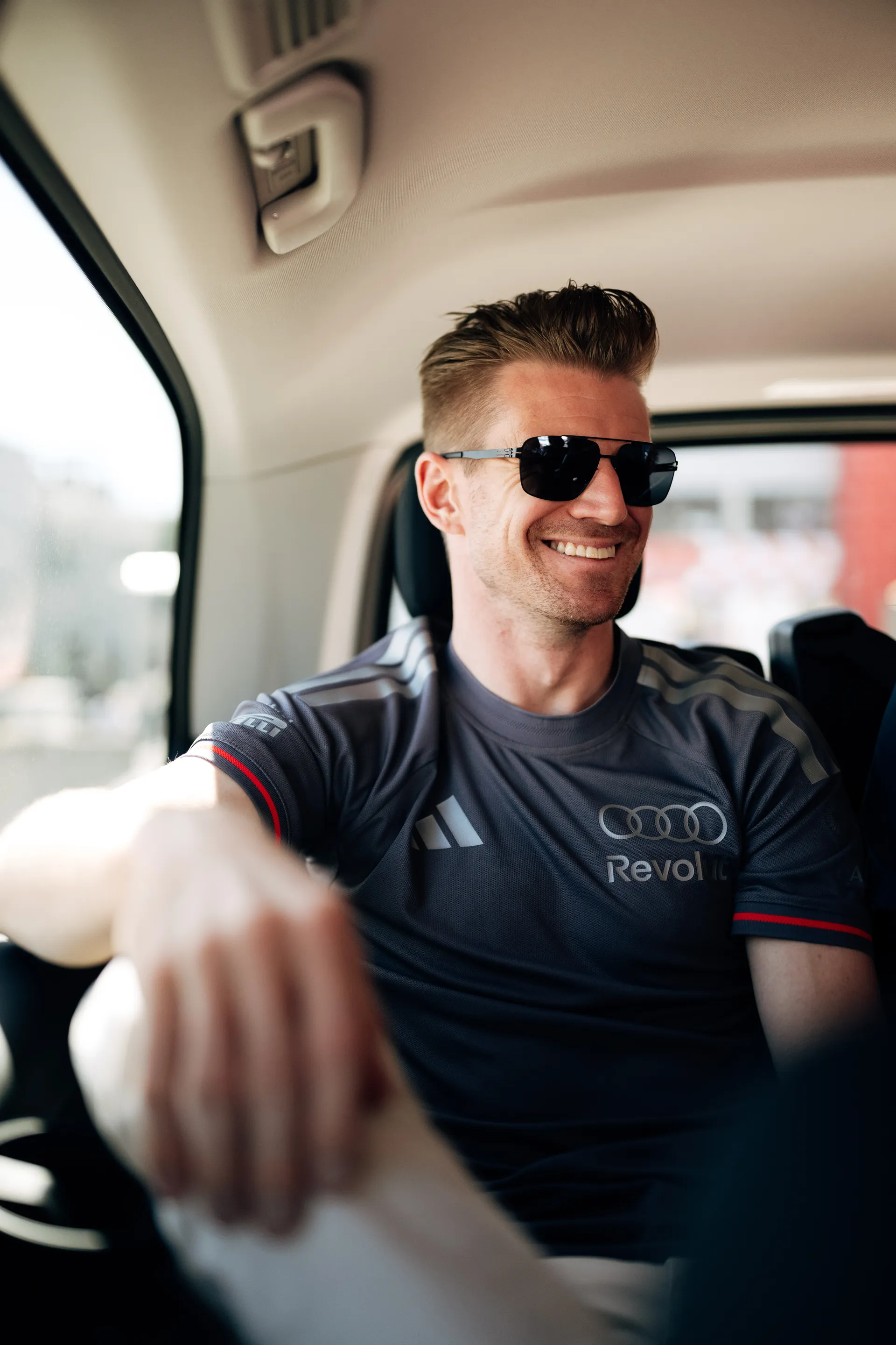 Nico Hulkenberg sitting inside a car, smiling and wearing sunglasses in Audi Revolut F1® Team apparel during Barcelona race weekend transport