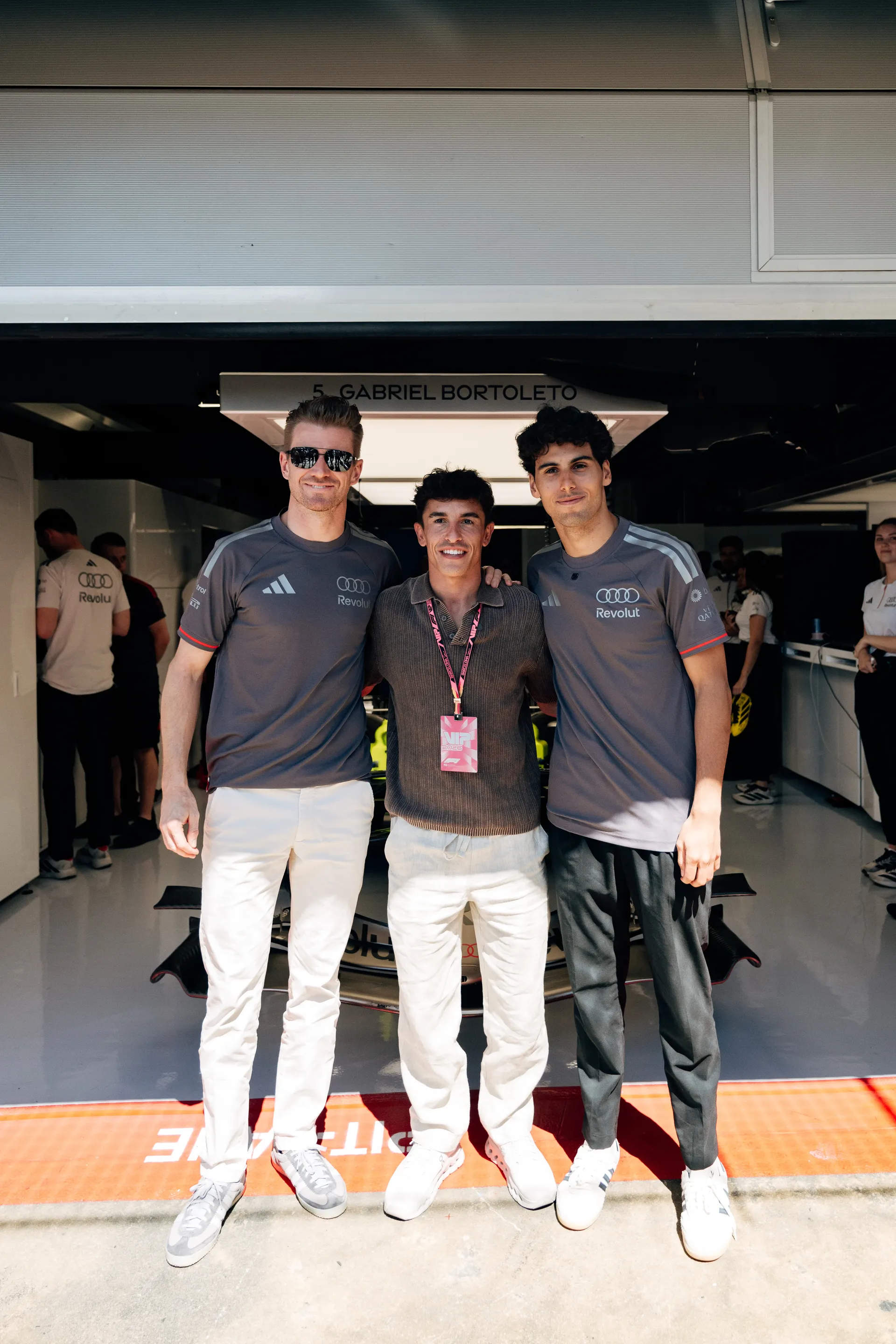 Nico Hulkenberg and Gabriel Bortoleto posing with a guest in front of the garage, with the Audi Revolut F1® Team car behind them