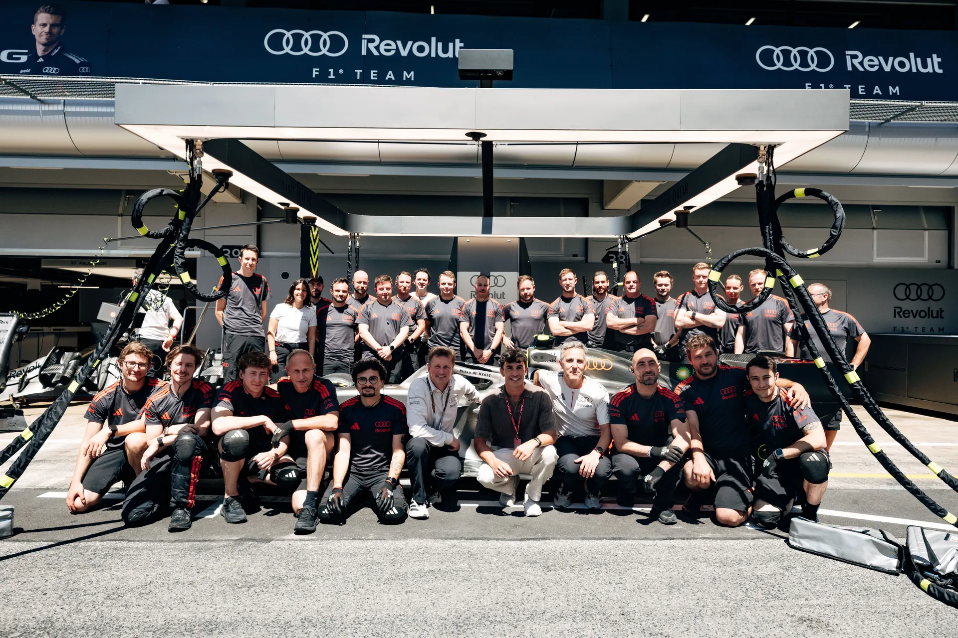 Full Audi Revolut F1® Team group photo in the pit lane with engineers, mechanics, and drivers gathered around the car