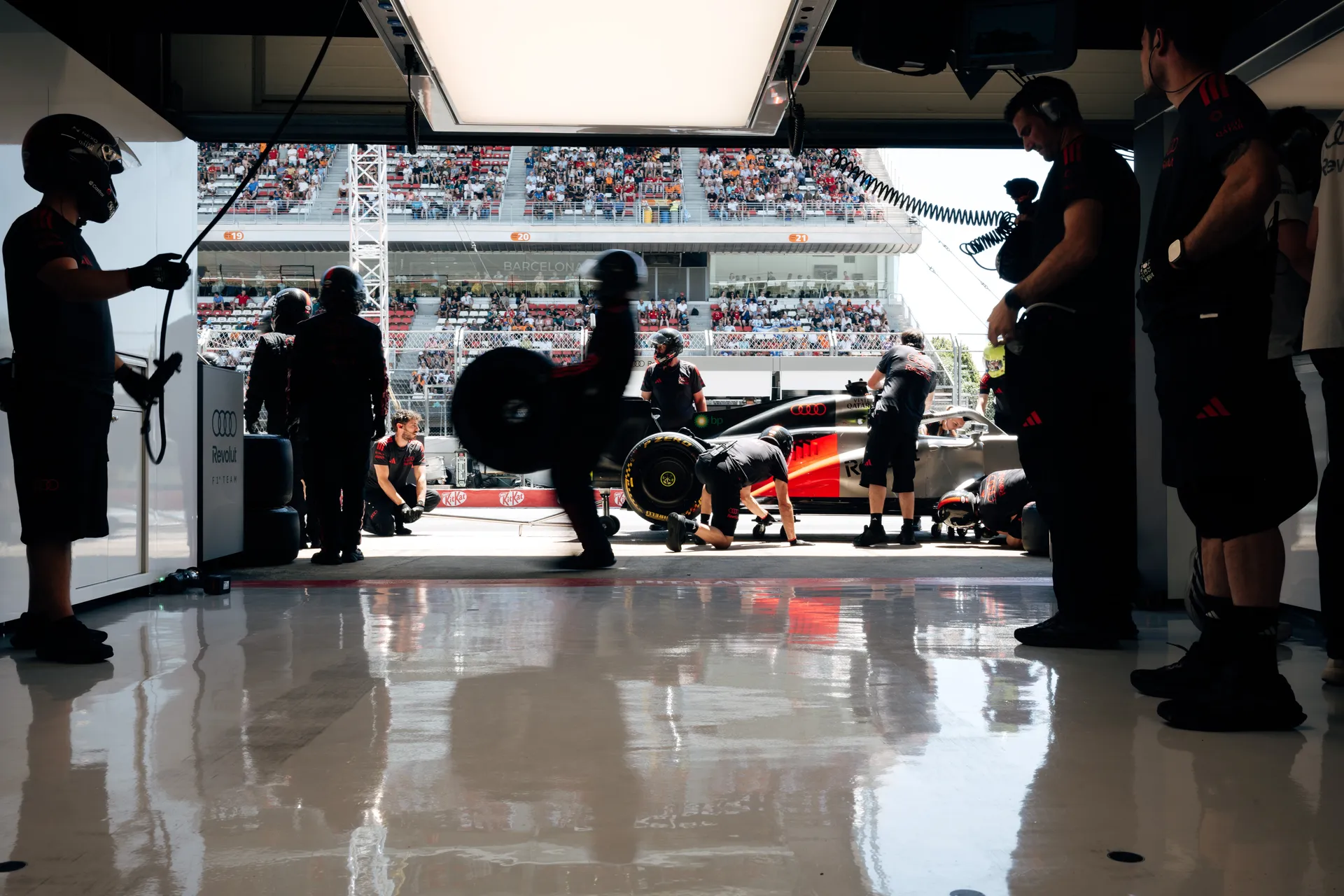 Audi Revolut F1® Team pit crew preparing a Formula One car inside the garage at Circuit de Barcelona-Catalunya, with mechanics carrying tires and a full grandstand visible outside