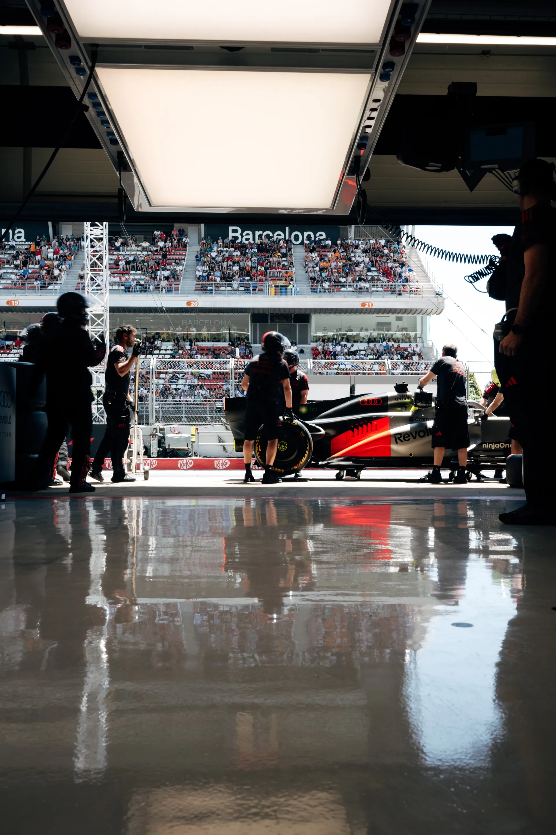 Audi Revolut F1® Team Formula One car positioned at the garage exit in Barcelona, surrounded by pit crew preparing for a session with the grandstand crowd in the background