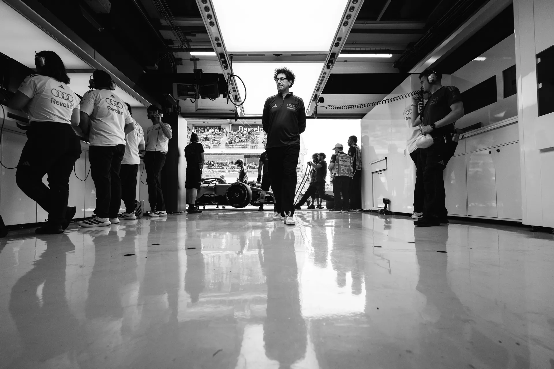 Black and white image of Mattia Binotto from Audi Revolut F1® Team engineer walking through the garage while team members work around the car in the background in Barcelona