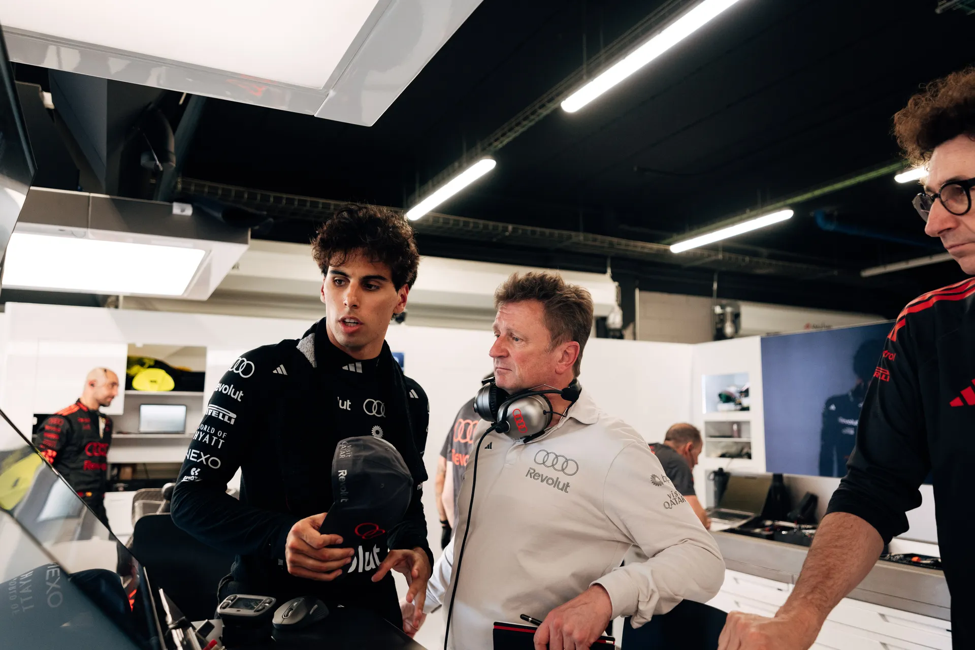 Gabriel Bortoleto discussing with Allan McNish from Audi Revolut F1® Team engineer inside the garage while holding a team cap, surrounded by technical equipment and crew