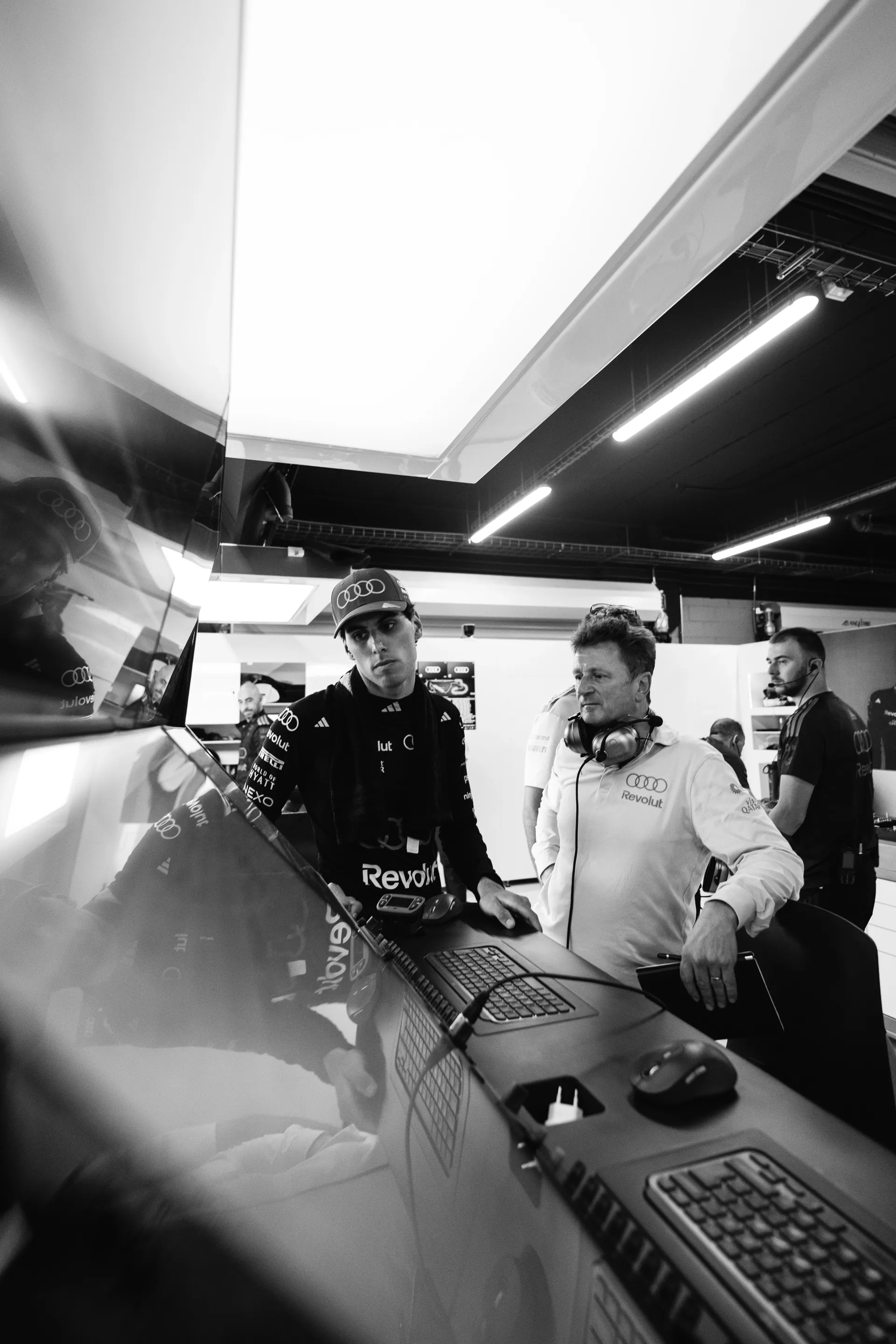 Black and white image of Gabriel Bortoleto reviewing data on a screen with an Audi Revolut F1® Allan McNish wearing a headset inside the garage