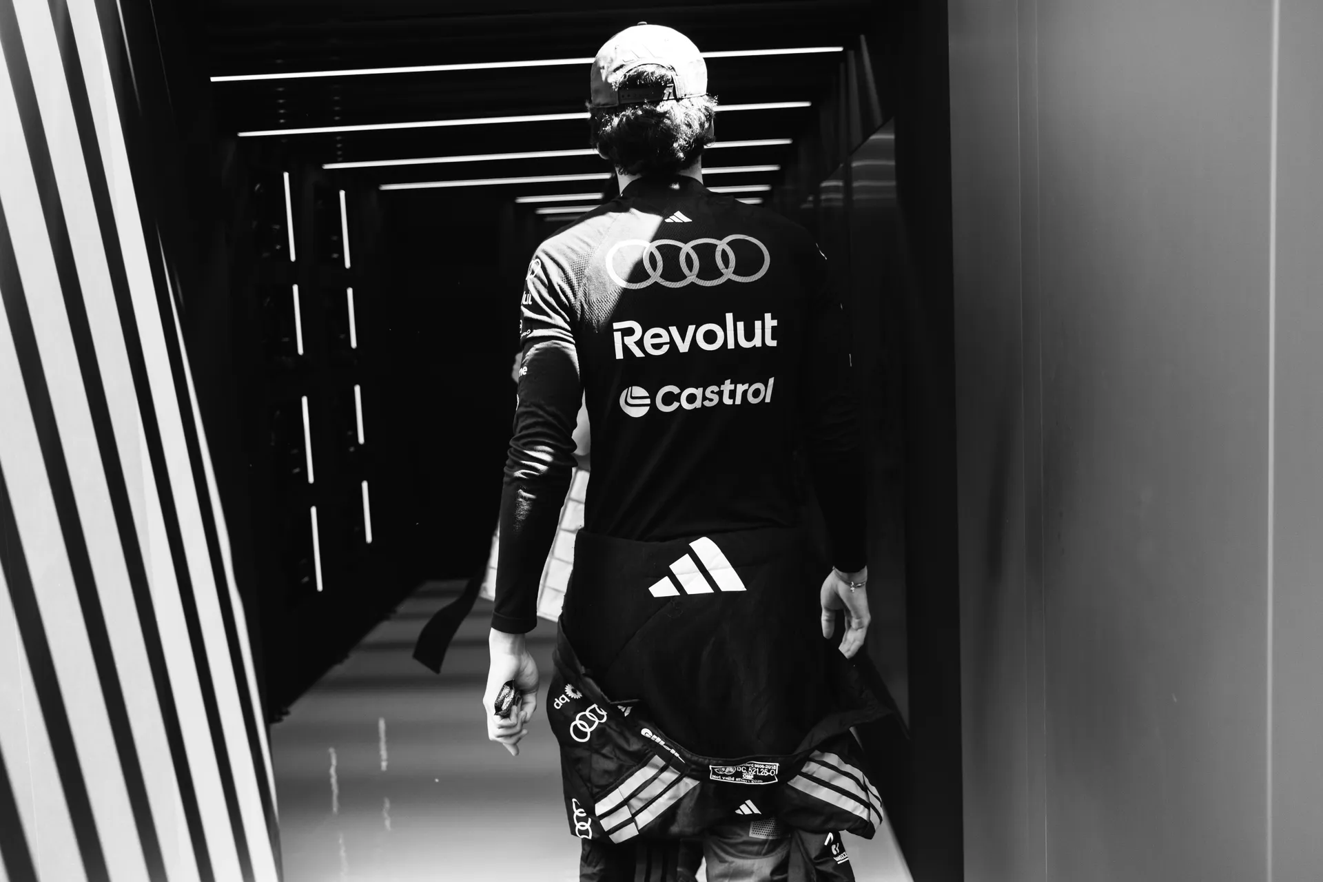 Audi Revolut F1® Team driver Gabriel Bortoleto walking through a narrow paddock corridor from behind, wearing full team kit with Audi, Revolut, and Castrol branding
