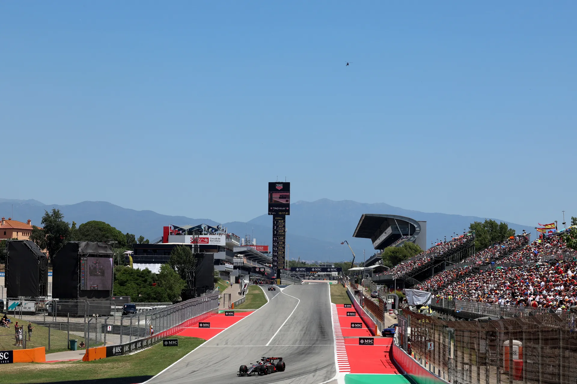 Wide shot of the Circuit de Barcelona-Catalunya main straight, with an Audi Revolut F1® Team car accelerating uphill past the pit lane as packed grandstands and the timing tower frame the scene.