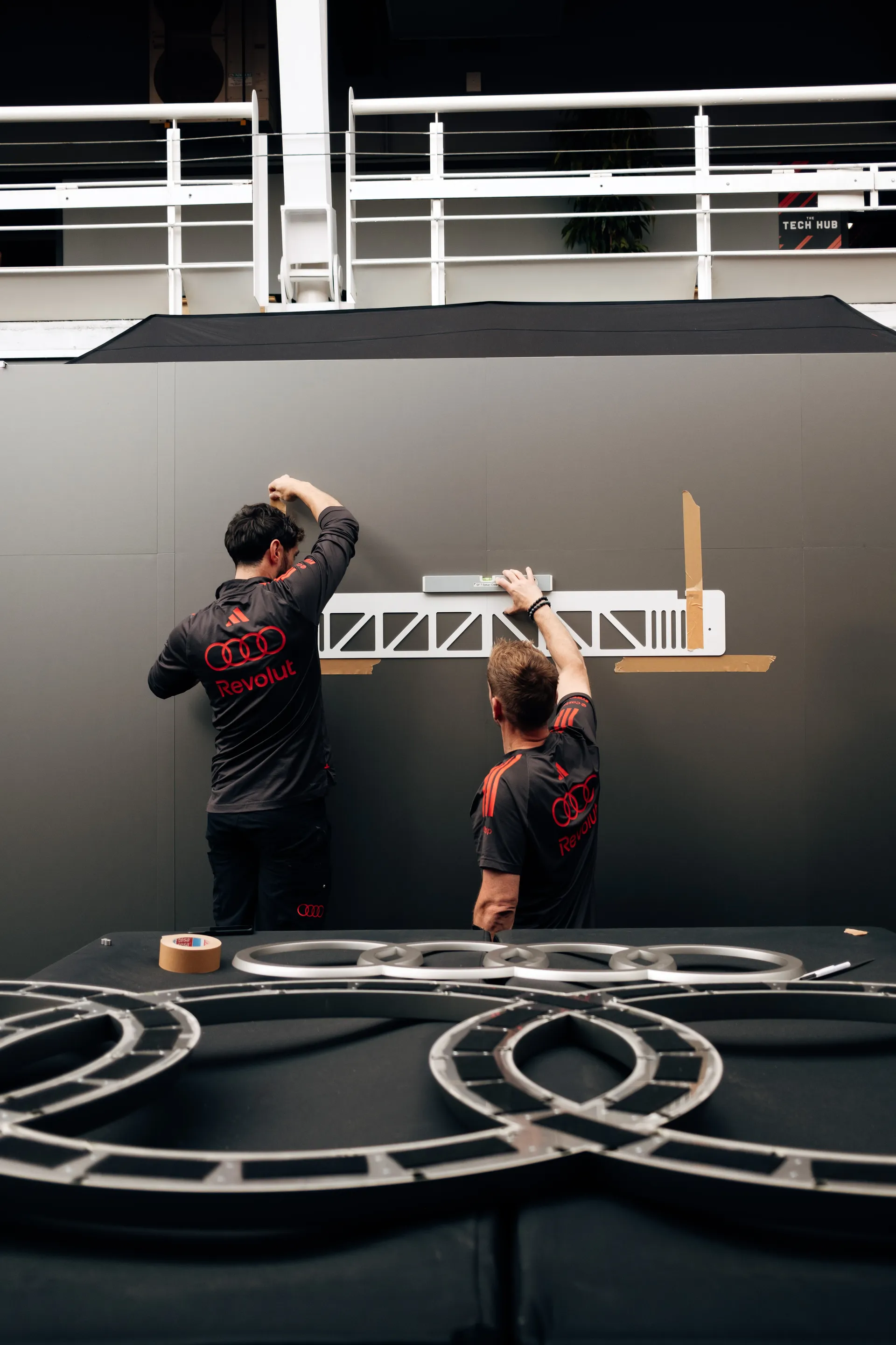 Two Audi Revolut F1® Team crew members align and mount a technical panel inside the garage, using measuring tools and tape for precision installation.