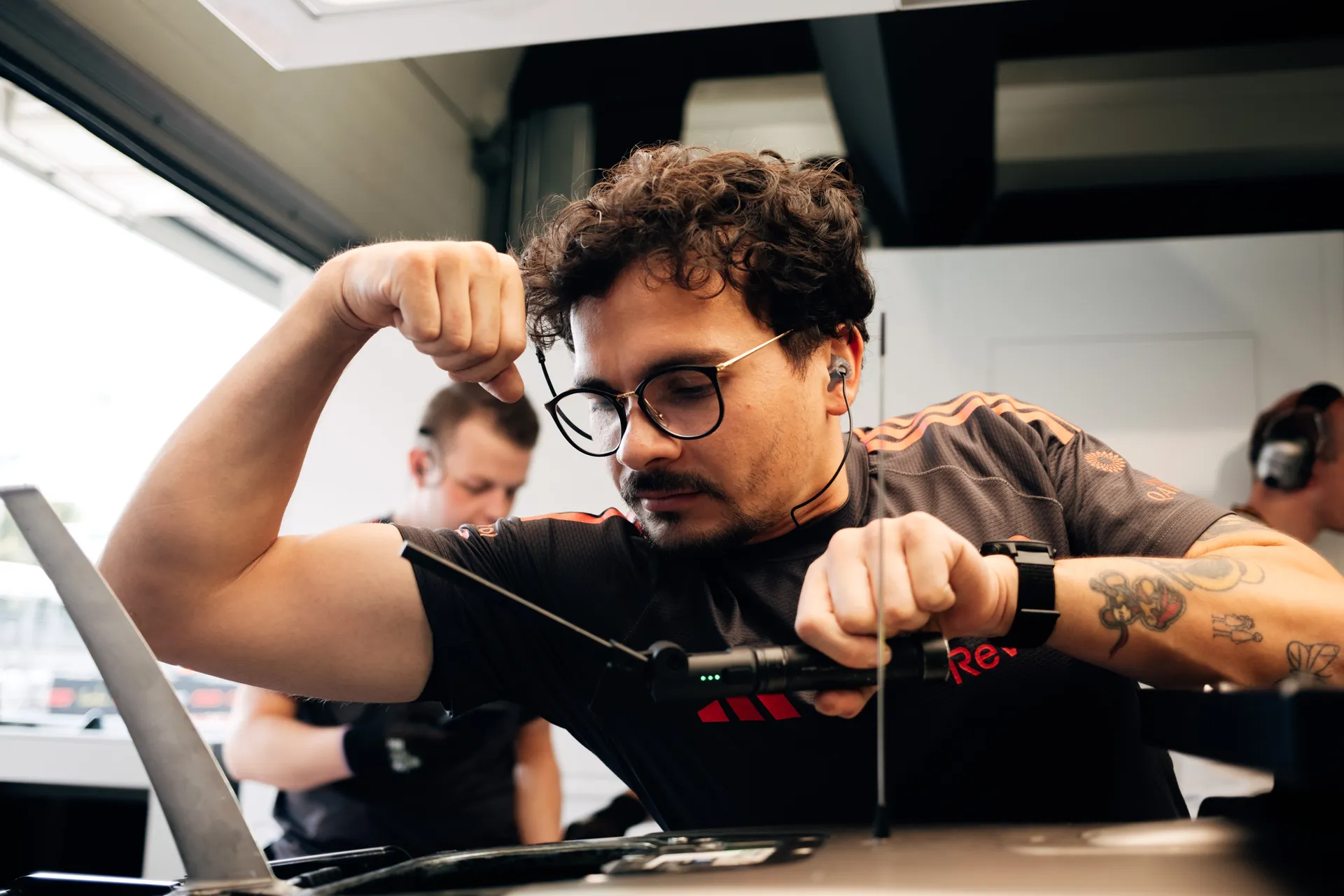 Audi Revolut F1® Team mechanic adjusts a component with a handheld tool, focusing intensely while wearing glasses and in-ear communication.