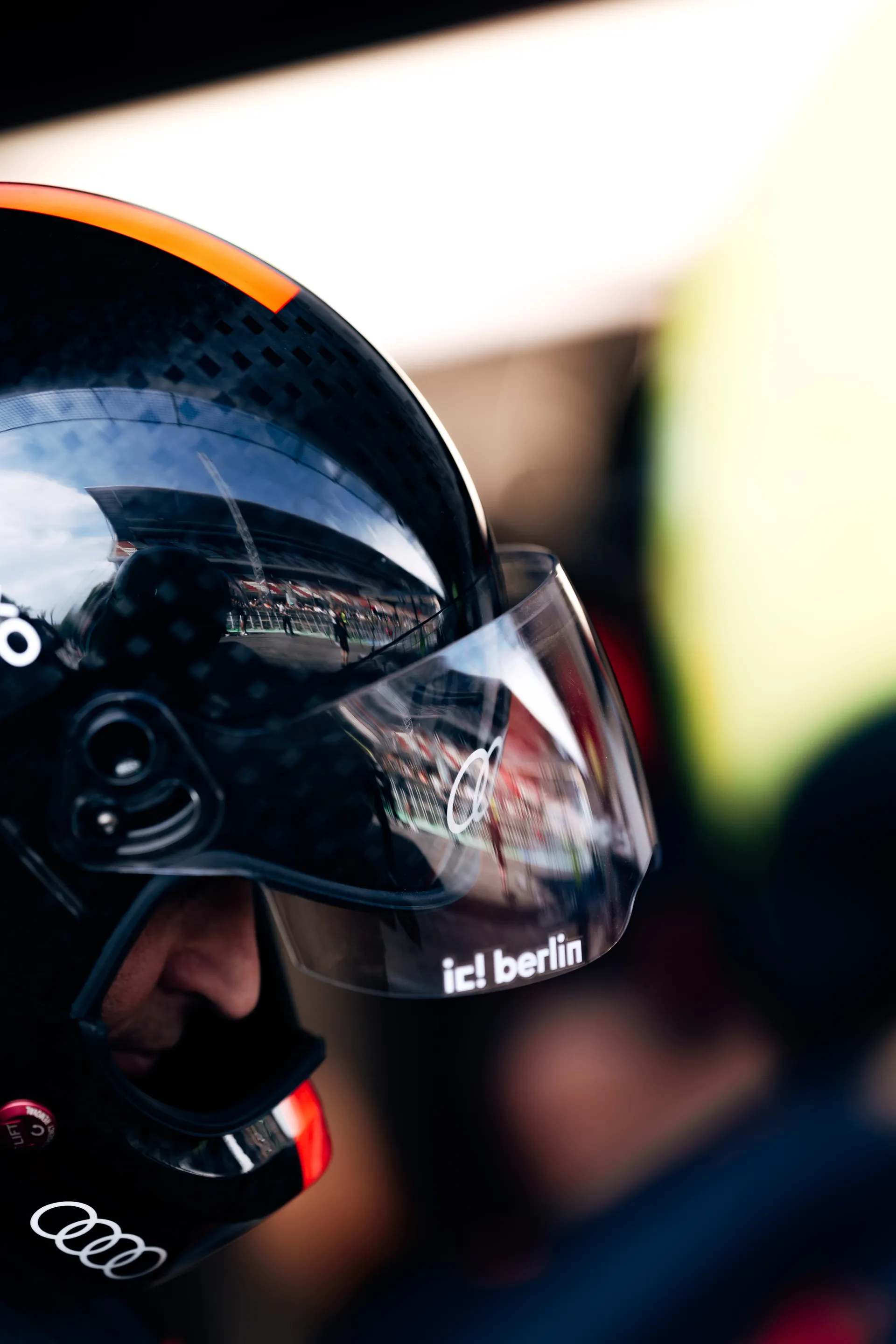 Close-up of a black Audi helmet with visor reflecting the pit lane and grandstand environment.