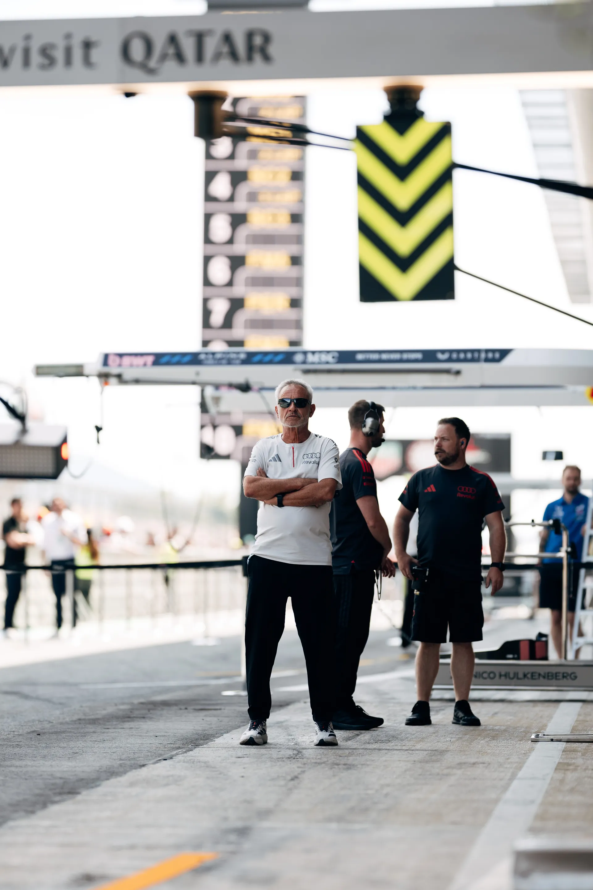 Audi Revolut F1® Team personnel stand in the pit lane observing track activity, with Nico Hulkenberg’s pit area visible in the background.