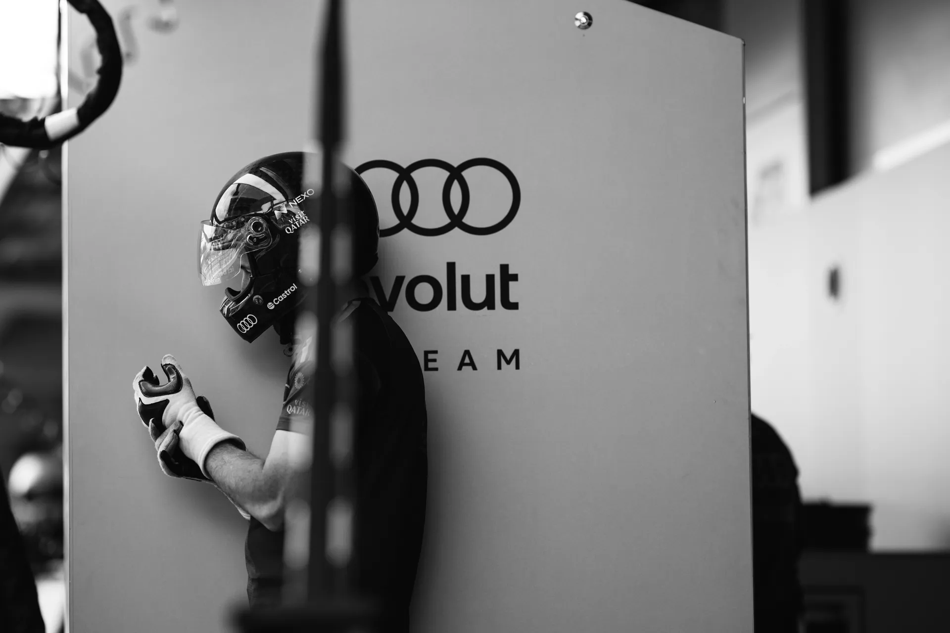 Black and white image of a helmeted Audi Revolut F1® Team crew member standing beside branded garage wall.