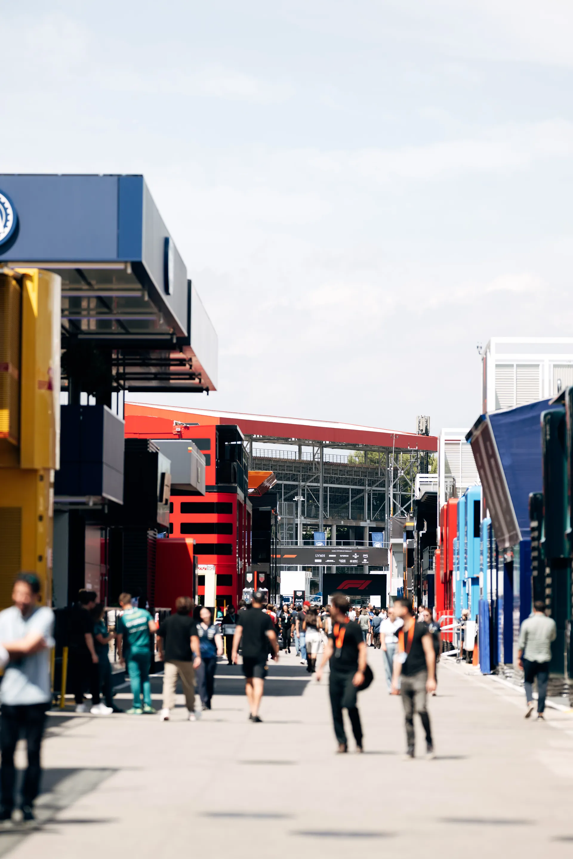 Wide paddock shot showing team hospitality units and personnel moving between garages during a race weekend.