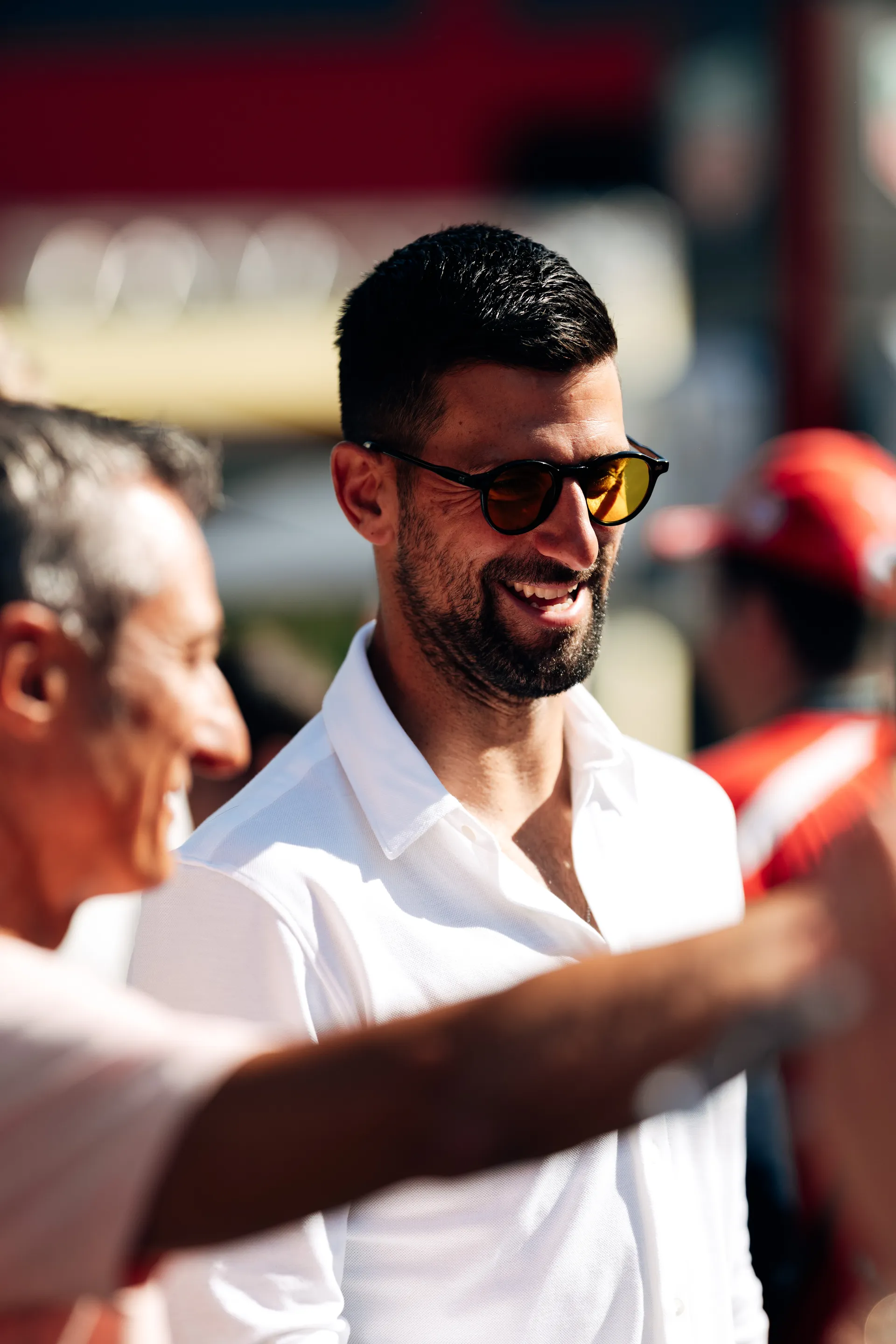 Novak Djokovic smiling in conversation in the paddock, wearing a white shirt and sunglasses