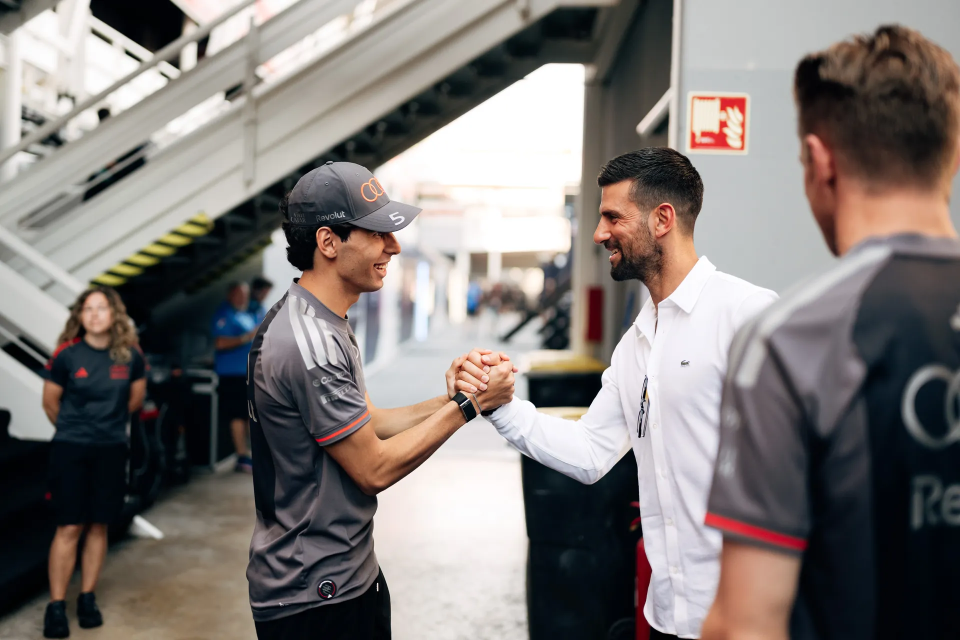 Gabriel Bortoleto and Novak Djokovic greeting each other with a handshake inside the Audi Revolut F1® Team garage