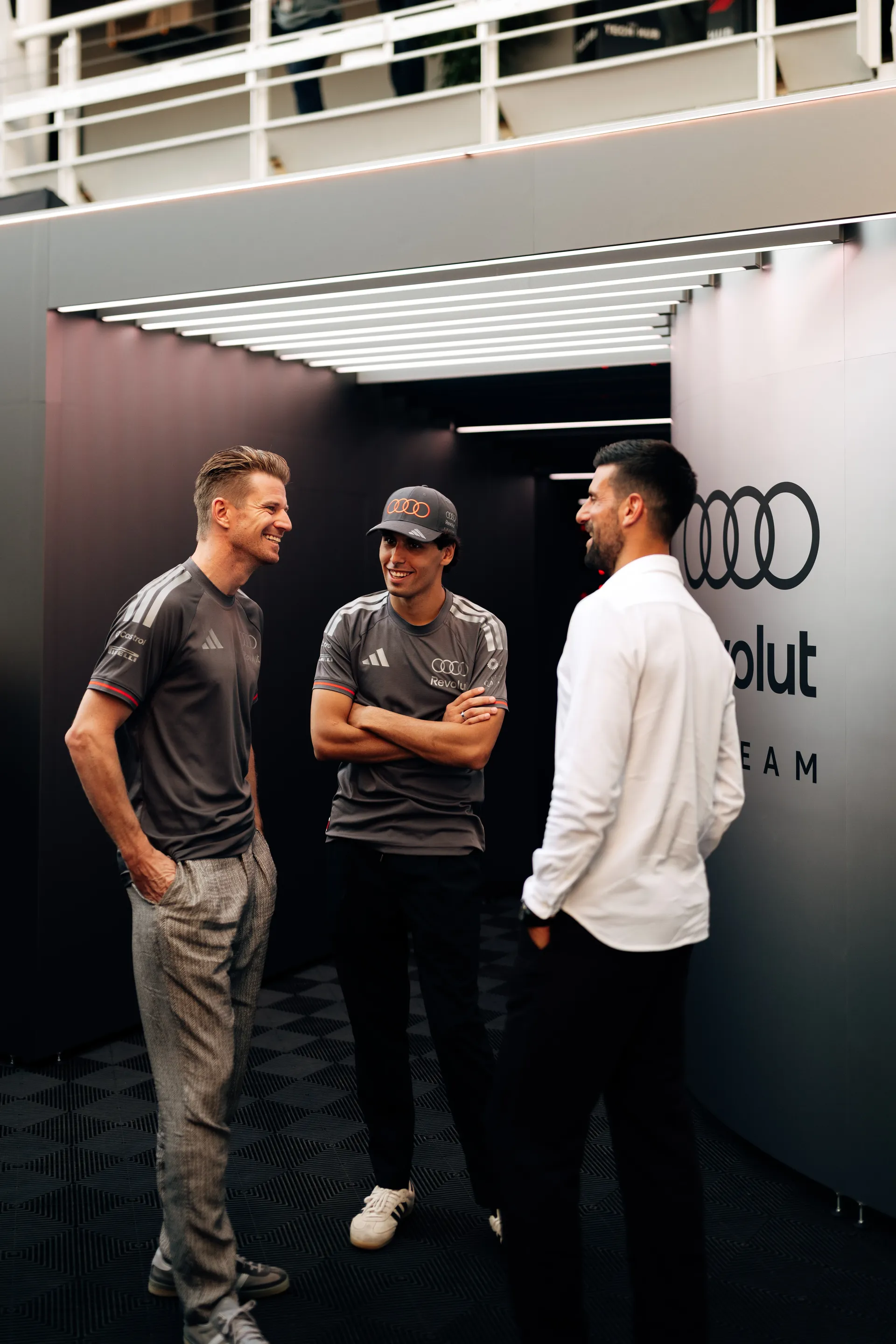 Nico Hulkenberg, Gabriel Bortoleto and Novak Djokovic sharing a relaxed moment and smiling inside the team garage
