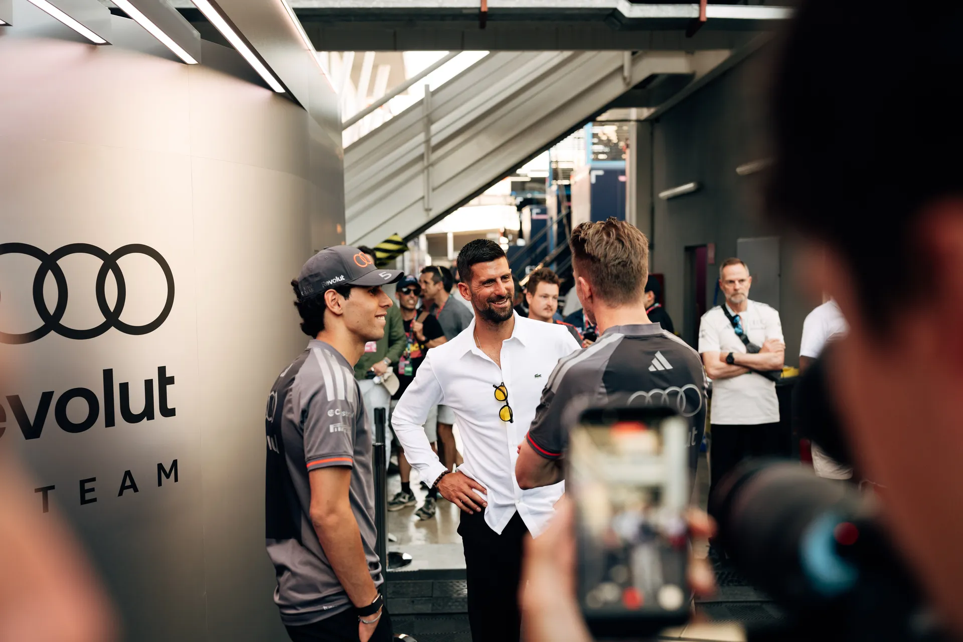 Novak Djokovic standing in the garage while Gabriel Bortoleto gestures towards equipment in the background