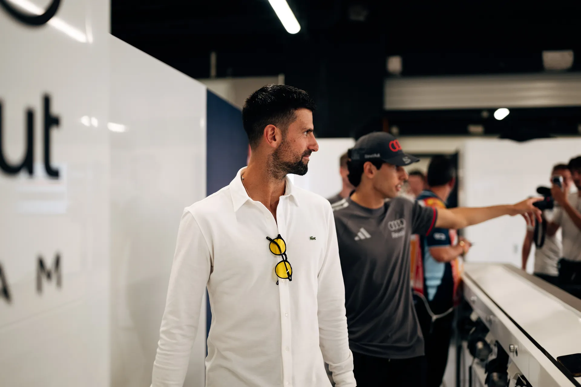 Gabriel Bortoleto and Novak Djokovic inspecting a Formula 1 steering wheel together in the pit lane
