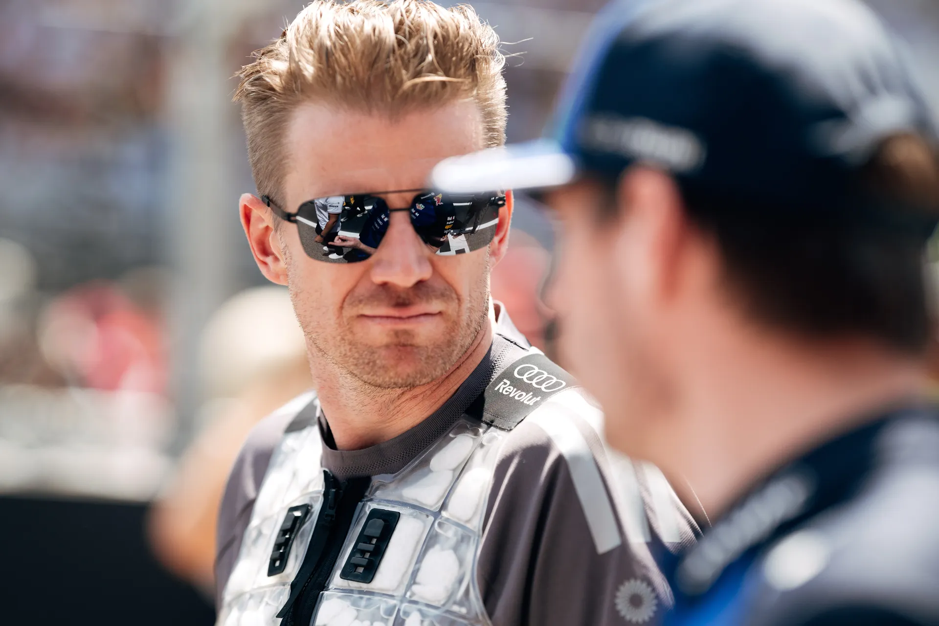 Nico Hulkenberg wearing sunglasses and a cooling vest, speaking to another driver during a paddock moment at the Barcelona Grand Prix