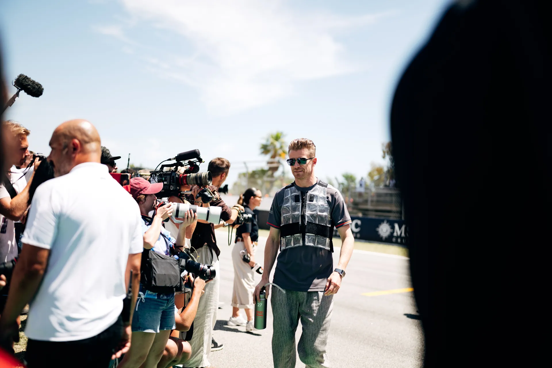 Nico Hulkenberg walking past photographers on the grid wearing a cooling vest, surrounded by media capturing pre-race moments