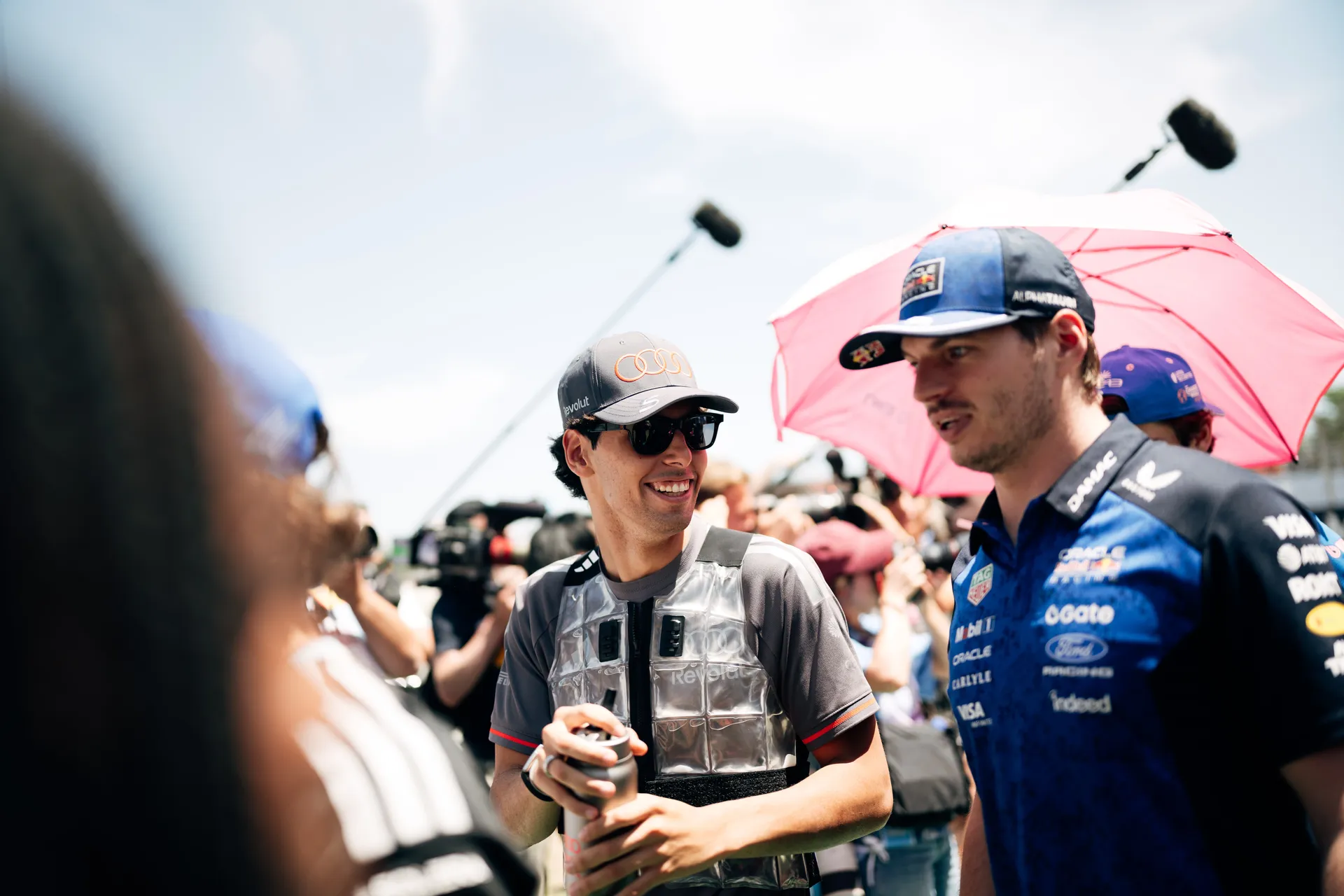 Gabriel Bortoleto smiling and talking with another driver under a sun umbrella during a relaxed pre-race grid interaction