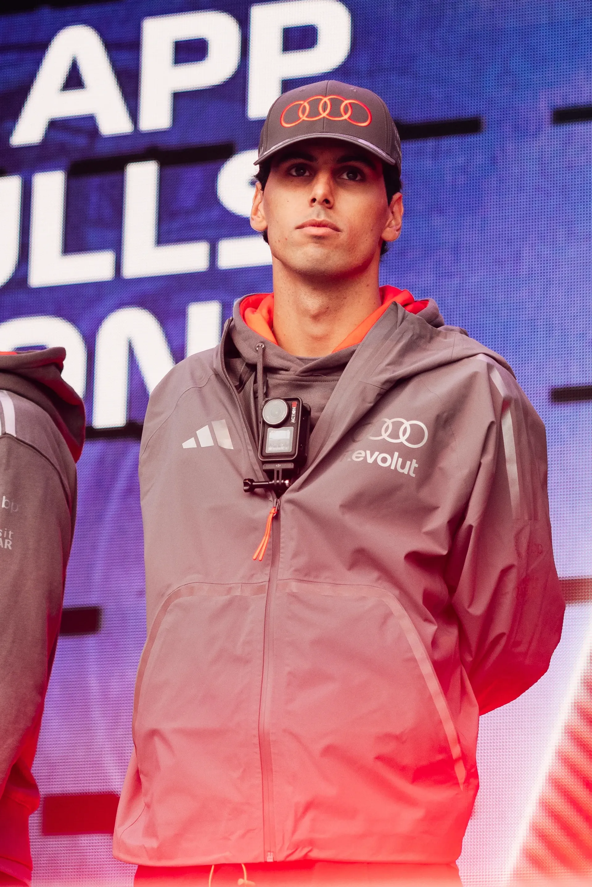Gabriel Bortoleto stands on stage at the Belgian Grand Prix fan forum, wearing Audi Revolut F1® Team gear with a camera mounted on his chest.