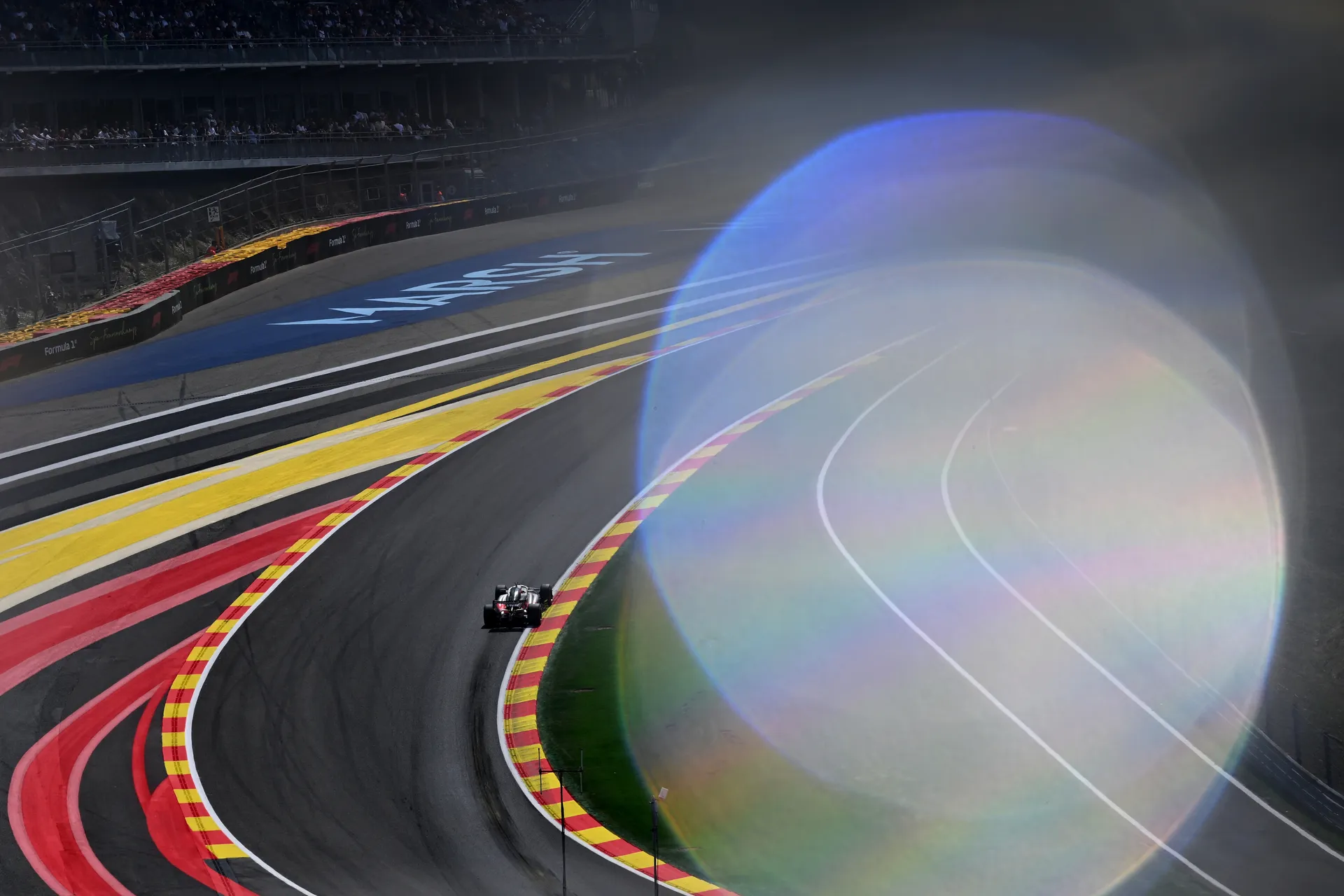 Formula 1 car driving through a sweeping corner at Spa Francorchamps during qualifying, with grandstands in the background and a strong lens flare partially covering the frame