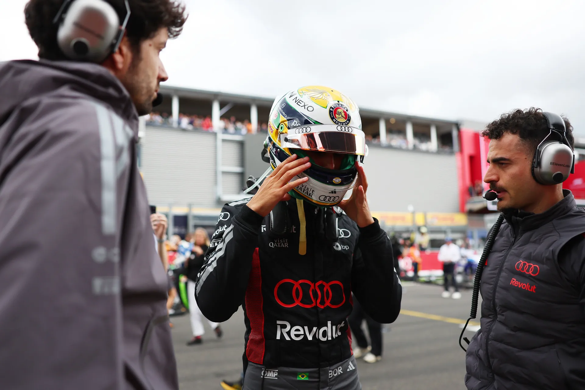 Gabriel Bortoleto adjusts his helmet on the grid while Audi Revolut F1® Team engineers stand alongside him at Spa-Francorchamps.