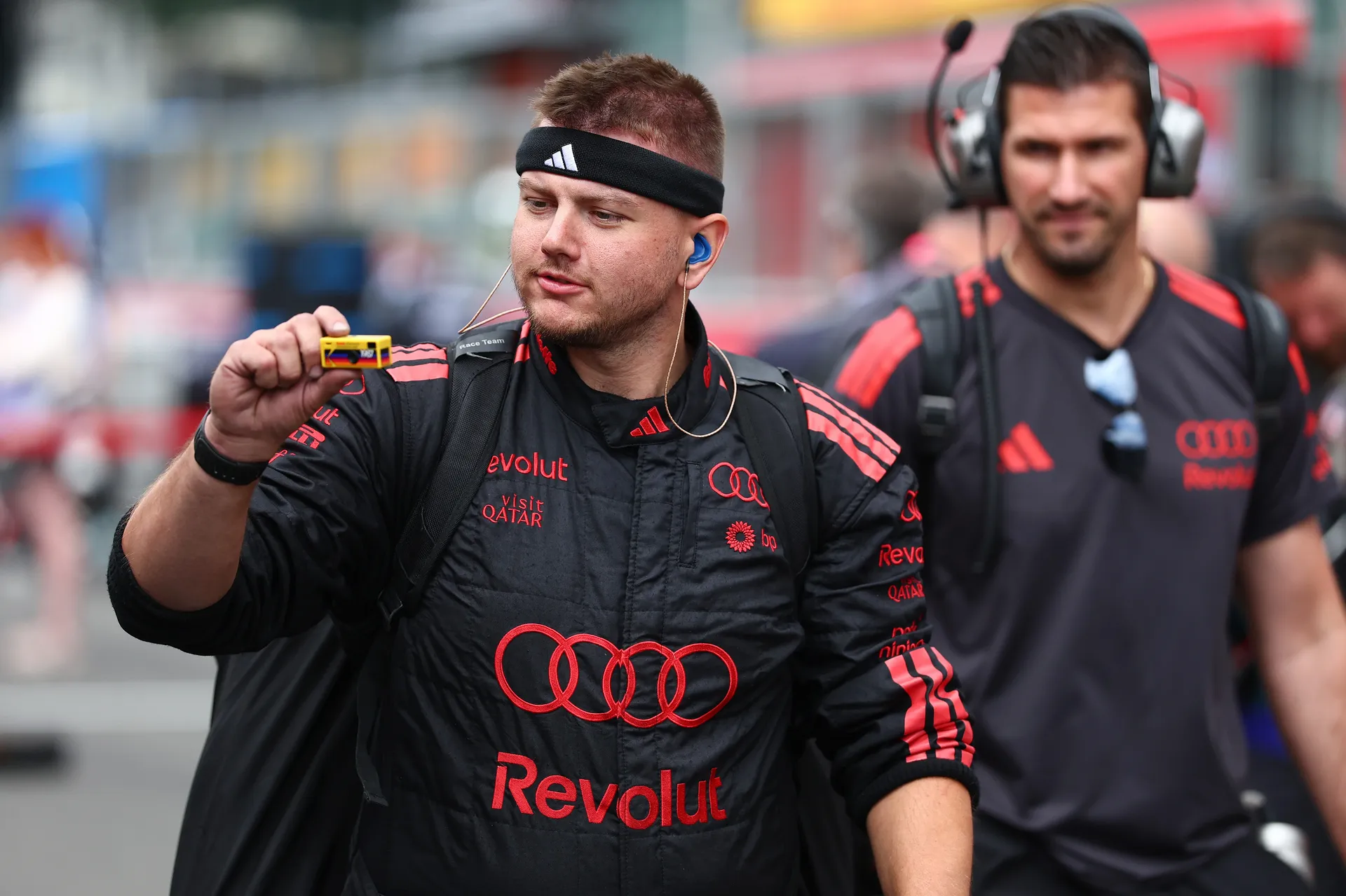 An Audi Revolut F1® Team crew member walks through the paddock holding a small camera, with another team member following behind.