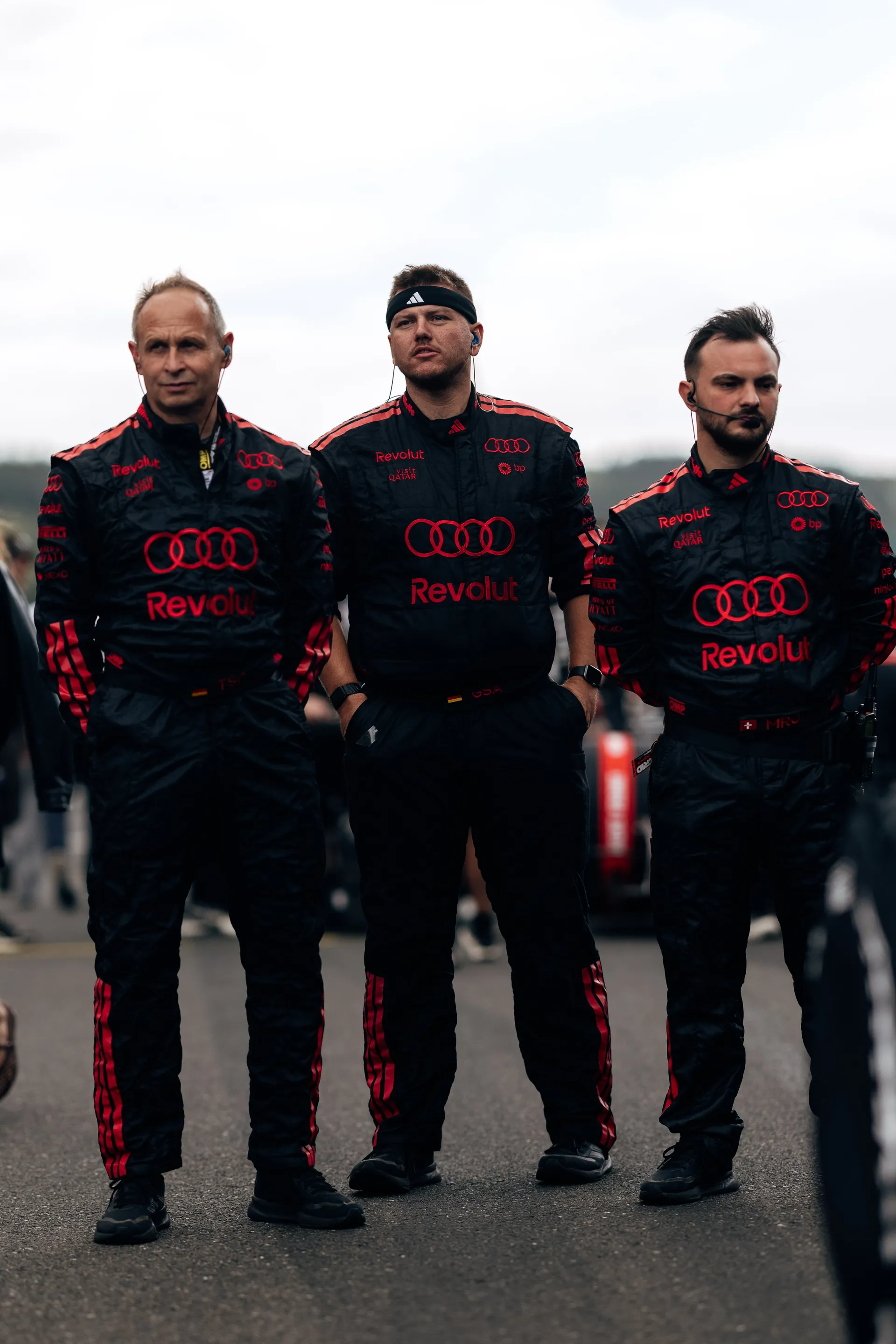 Three Audi Revolut F1® Team crew members stand side by side on the grid wearing race overalls and headsets during pre-race preparations at Spa-Francorchamps.