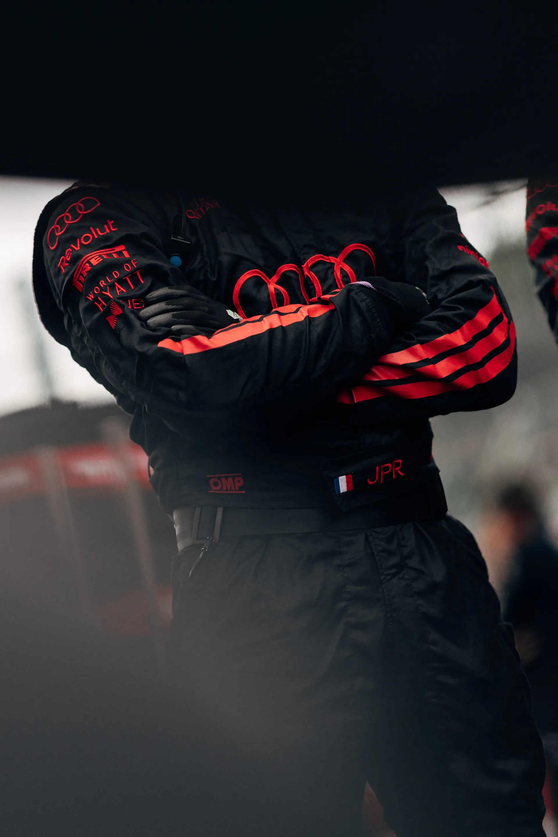 Audi Revolut F1® Team crew member stands with arms crossed in race overalls on the grid, partially framed in the foreground at Spa-Francorchamps.