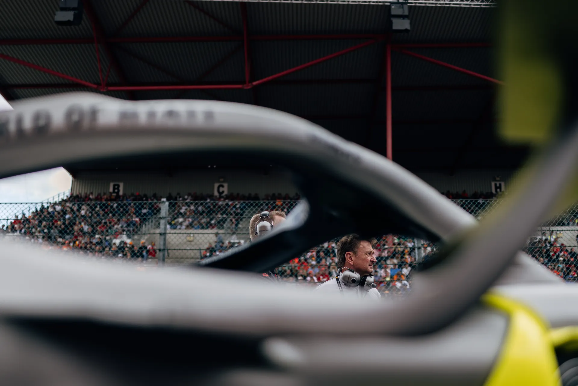 Allan McNish stands near the track in front of grandstands, seen through the structure of the Audi Revolut F1® Team car at Spa-Francorchamps.