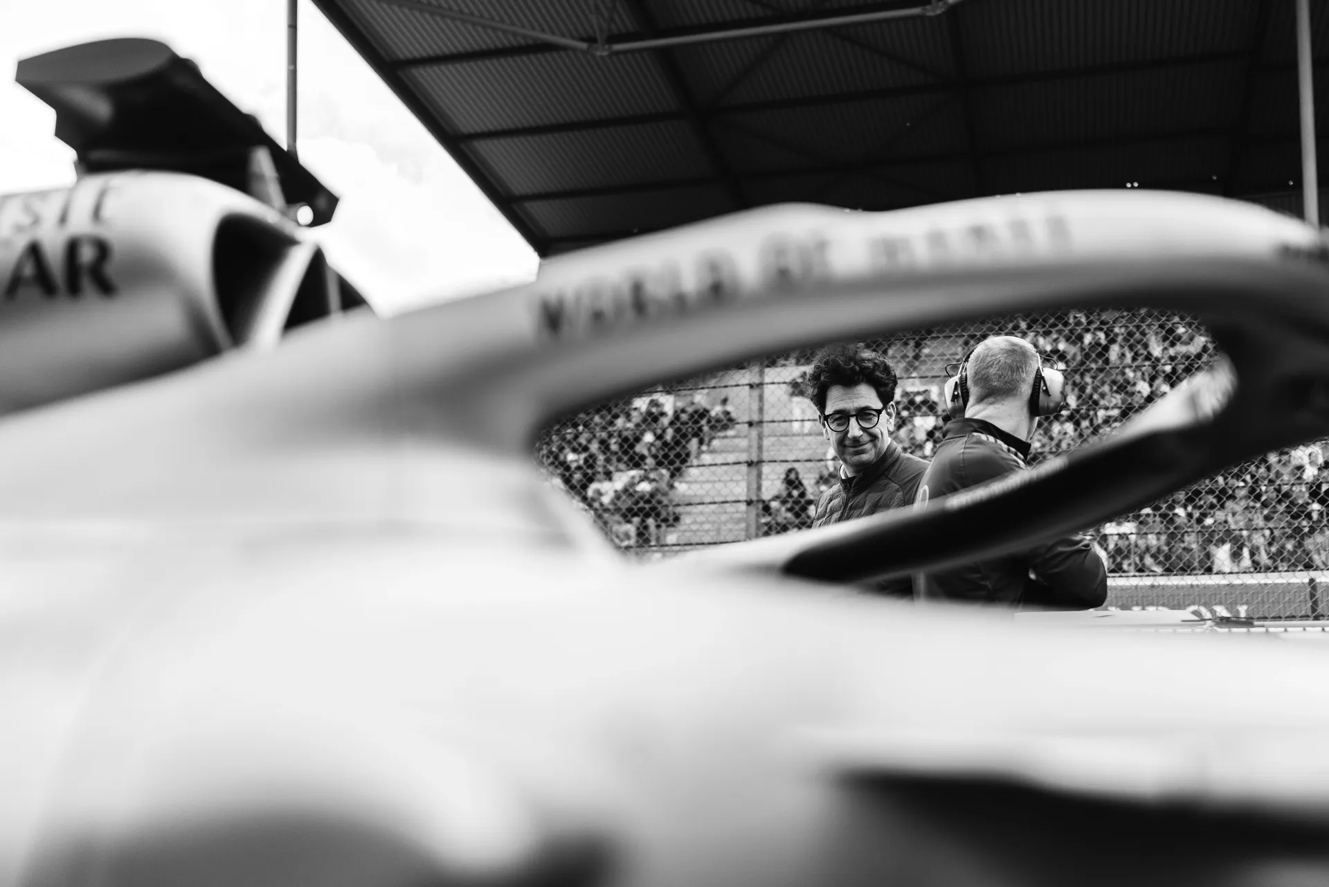 Black and white image of Mattia Binotto standing trackside with an Audi Revolut F1® Team colleague, framed through the halo of an Audi Revolut F1® Team R26 with grandstands in the background