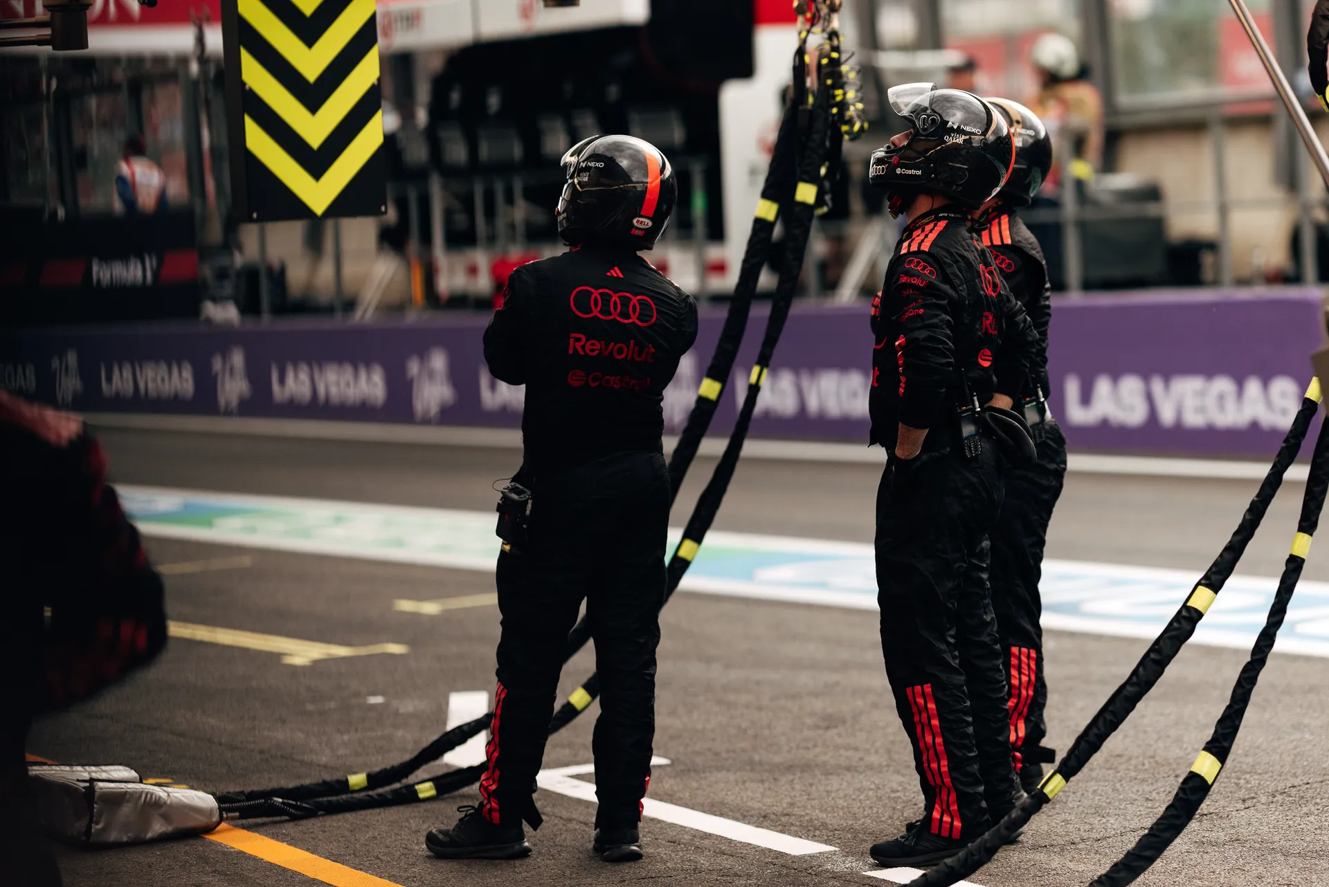 Three Audi Revolut F1® Team pit crew members in full race gear and helmets standing in the pit lane holding equipment hoses during race operations