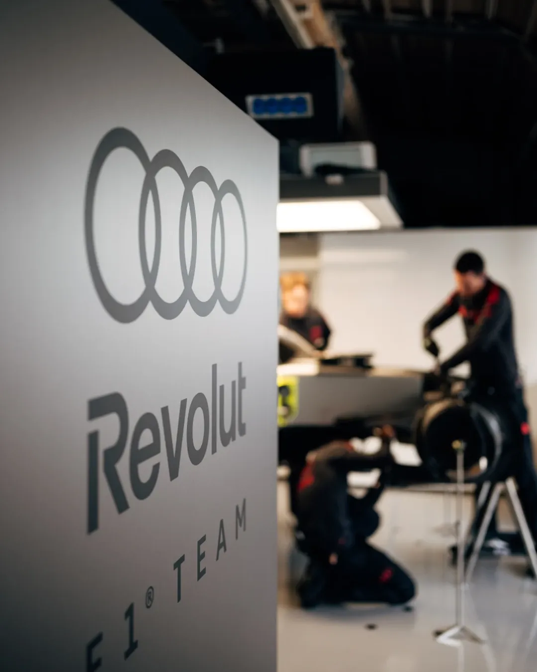 Audi Revolut F1® Team garage scene with mechanics working on the car, Audi rings and Revolut branding in the foreground.