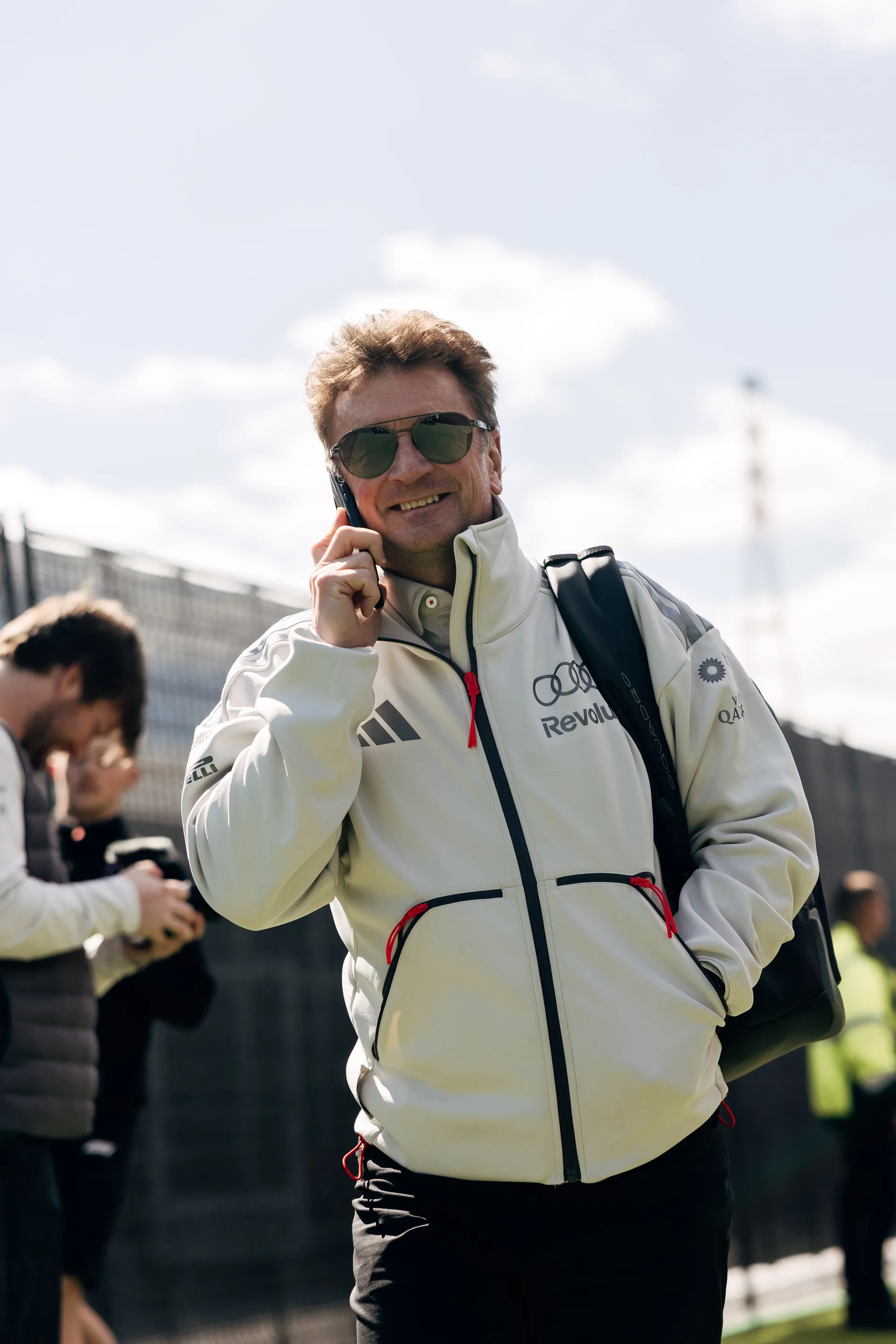 Allan McNish smiling while on the phone, wearing Audi Revolut F1® Team jacket in the paddock.