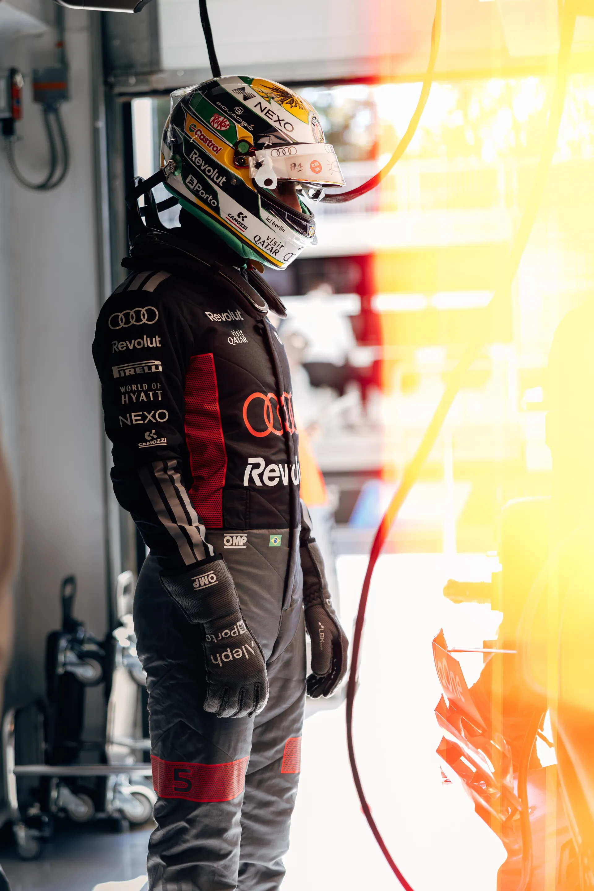 Gabriel Bortoleto in full race suit and helmet, standing in the garage with light flare.