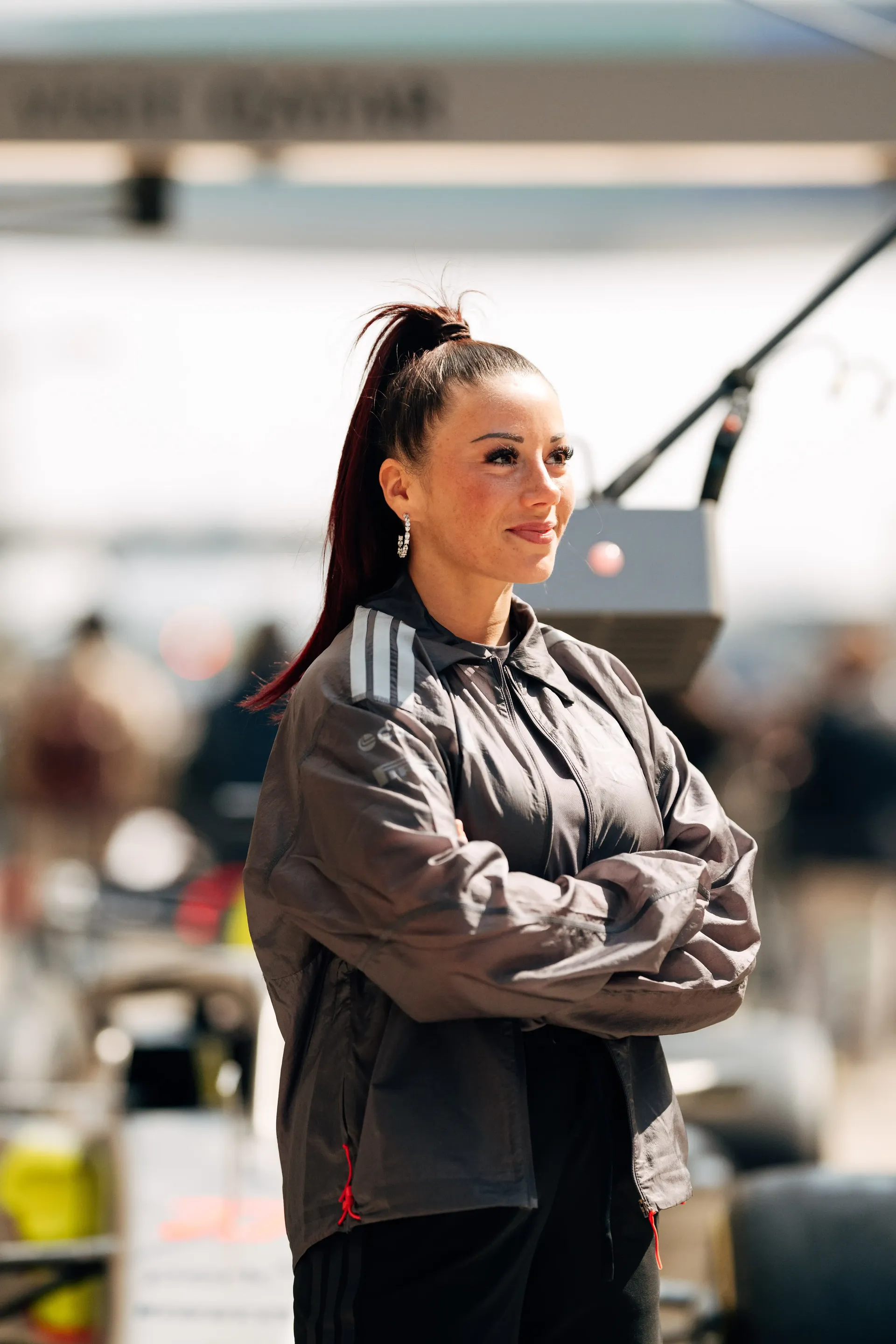Carrie Schreiner standing with arms folded, wider view showing race car and pit lane activity in China at Shanghai
