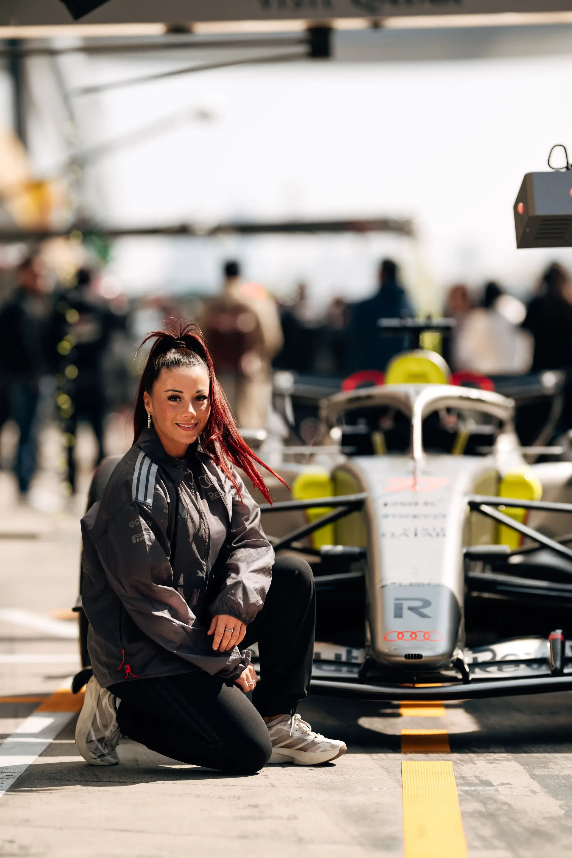 Carrie Schreiner kneeling beside race  car, looking at camera, pit lane crew blurred in background in China at Shanghai