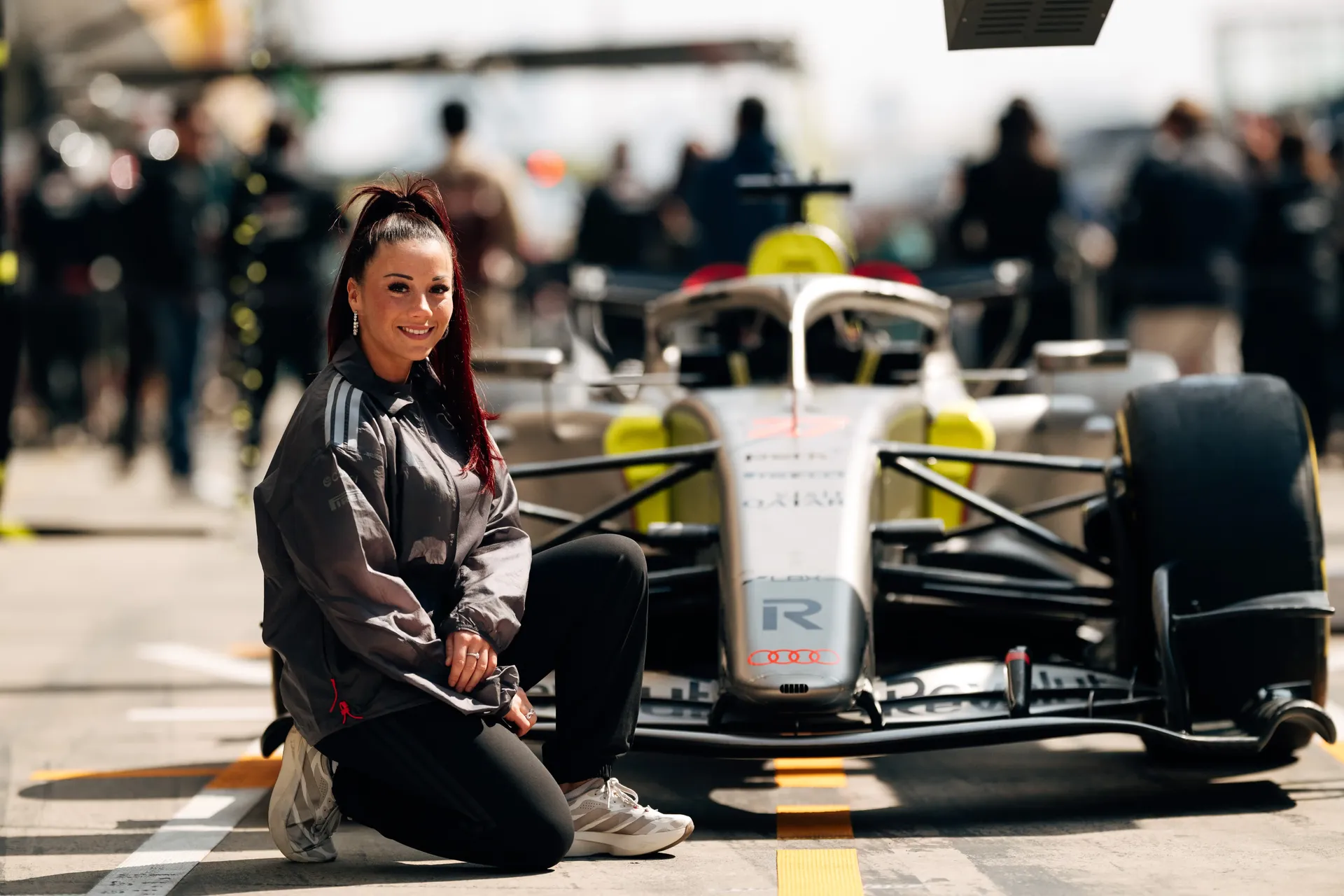 Carrie Schreiner kneeling beside race car, smiling at camera, pit lane activity behind in China at Shanghai