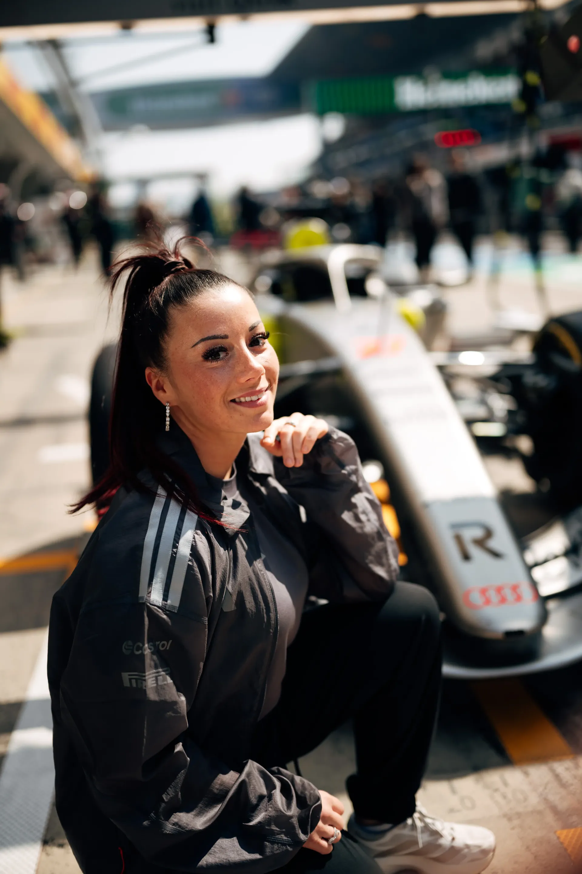 Carrie Schreiner squatting with one hand on chin, smiling, race car and pit lane in background in China at Shanghai