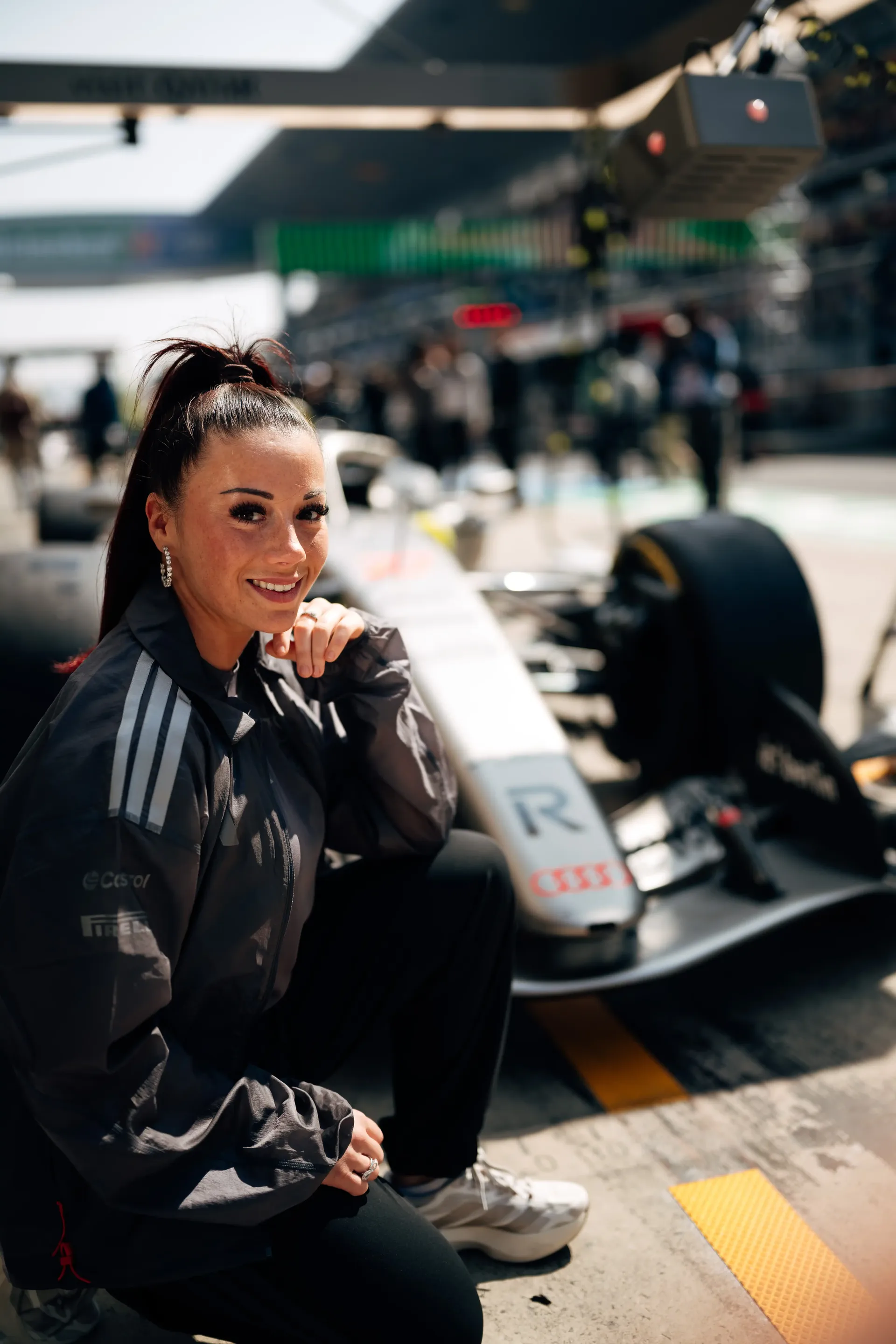 Carrie Schreiner squatting with one hand on chin, smiling, race car and pit lane in background in China at Shanghai