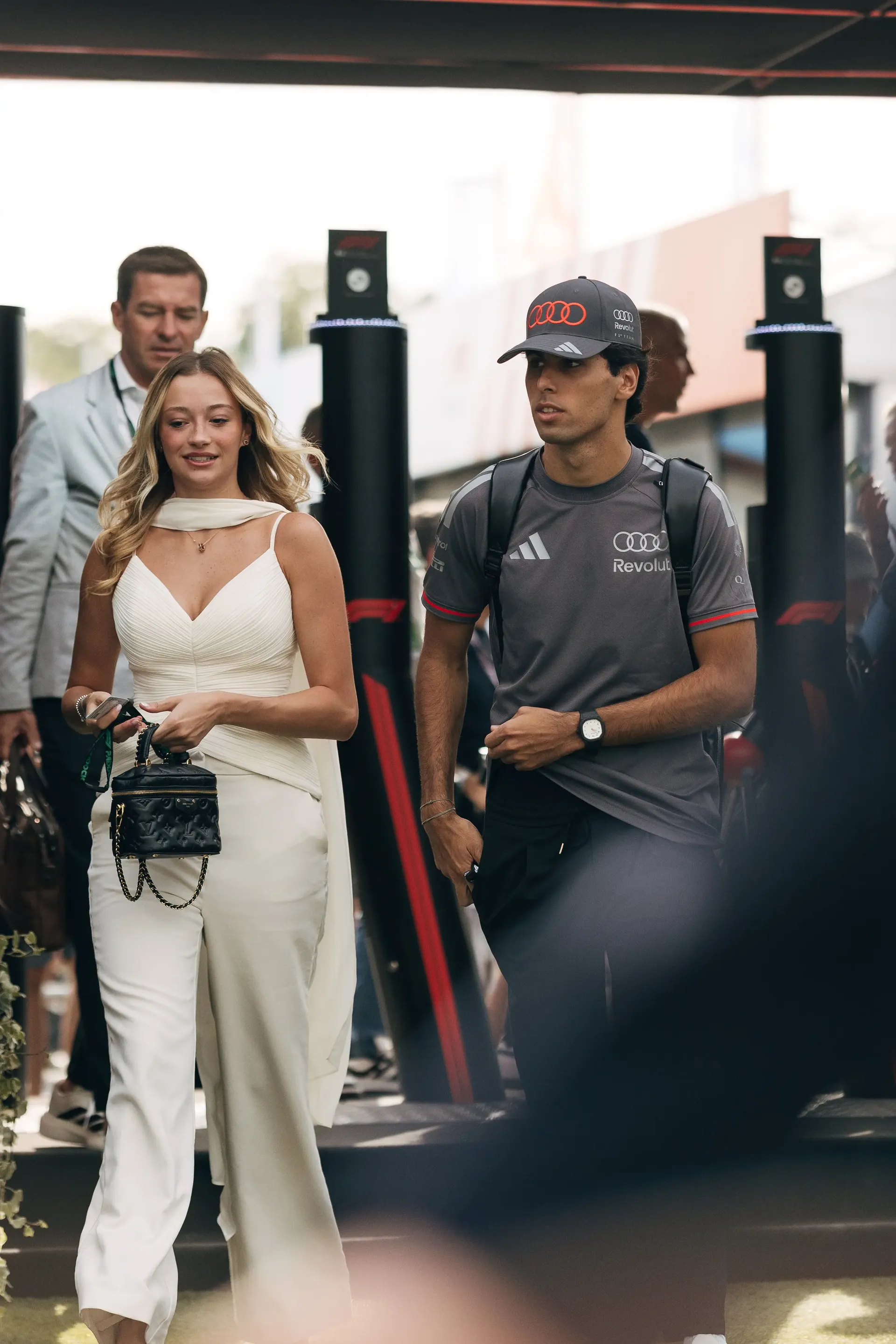 Audi Revolut F1® Team driver Gabriel Bortoleto walking through the paddock beside his girlfriend, with blurred foreground framing.