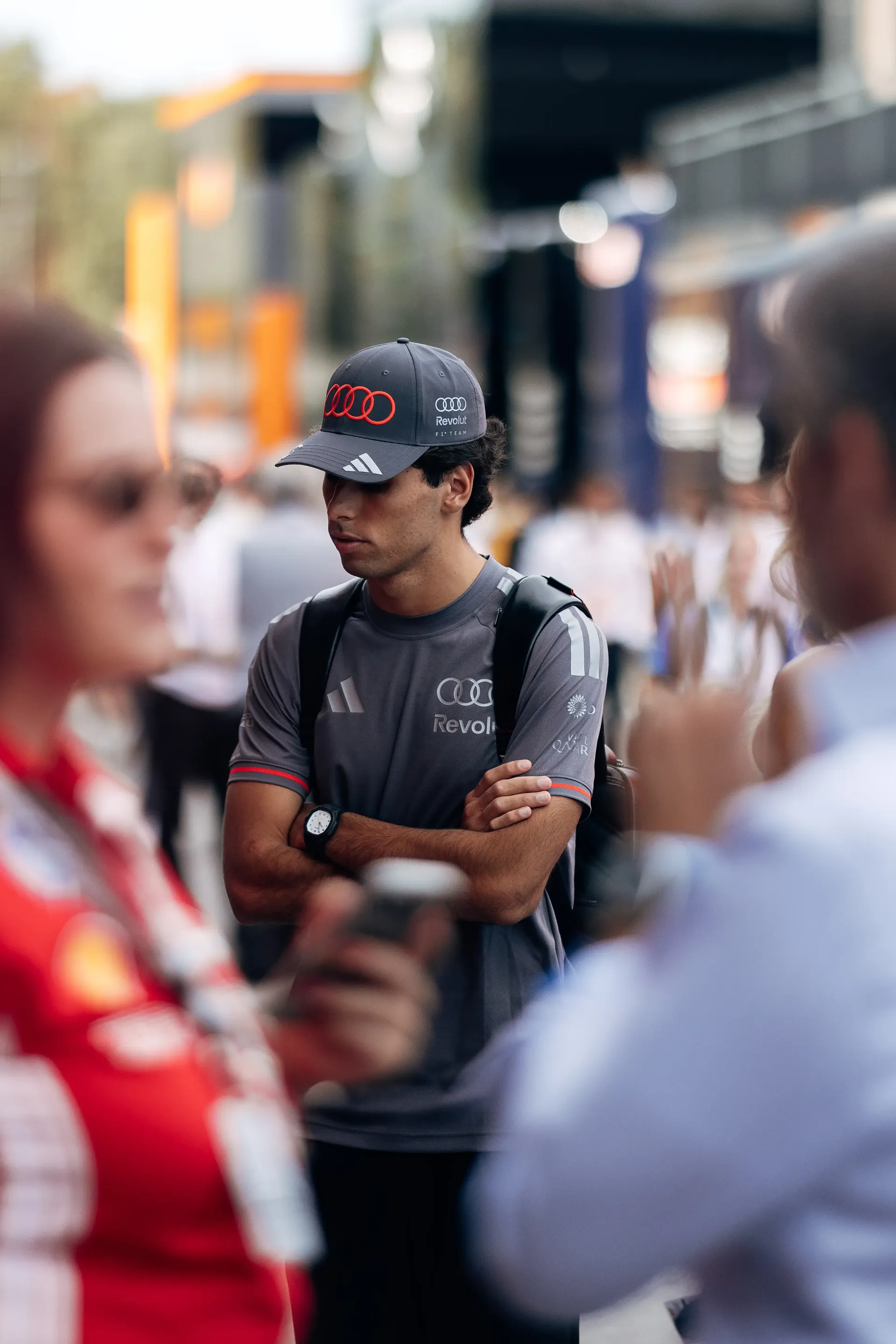 Audi Revolut F1® Team driver Gabriel Bortoleto standing with arms crossed in the paddock, surrounded by blurred figures.