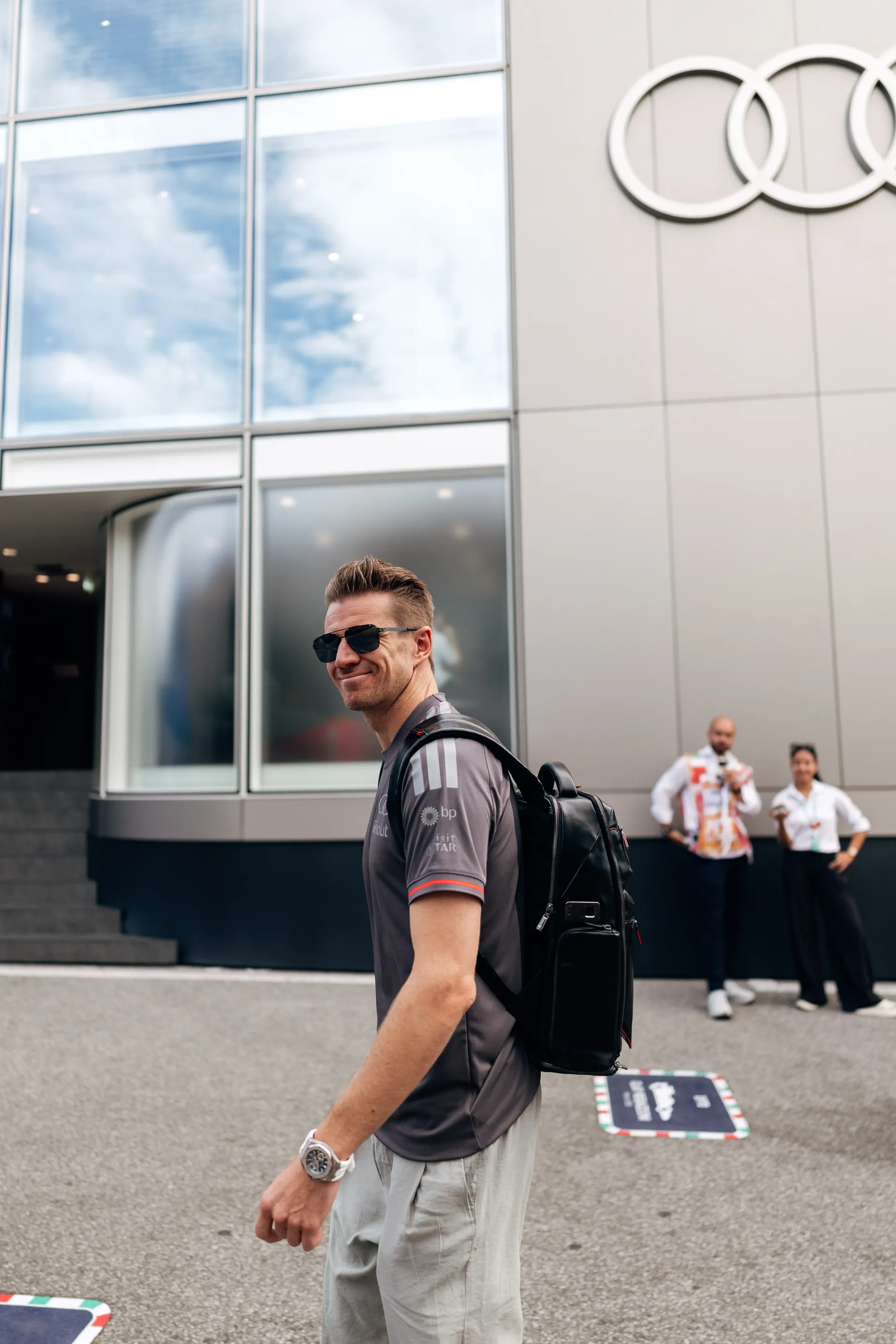 Audi Revolut F1® Team driver Nico Hulkenberg glancing back while walking outside the Audi Revolut F1 Team hospitality building with the Audi logo in the background.