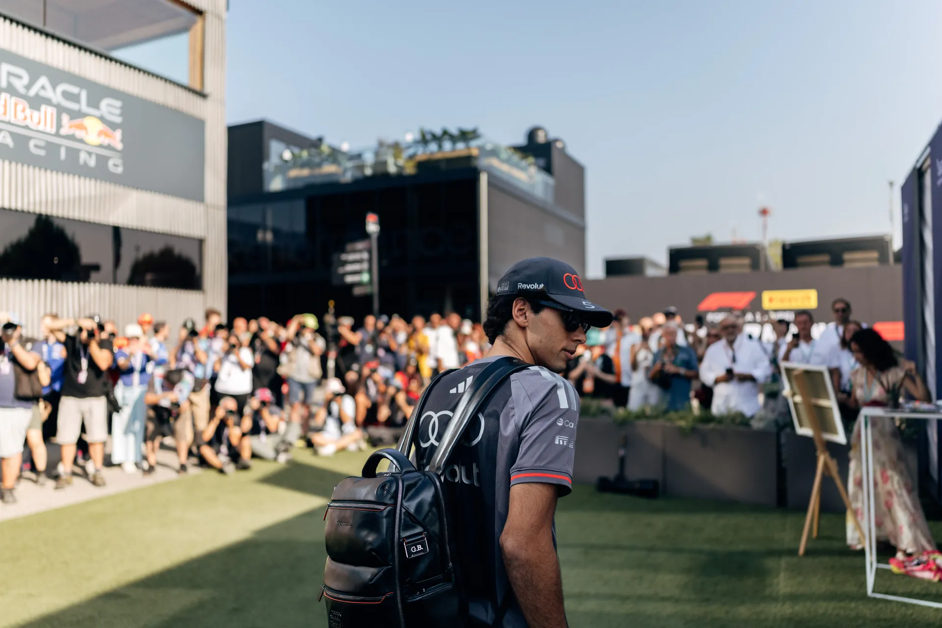 Gabriel Bortoleto, driver of the Audi Revolut F1® Team, walks through the Monza paddock carrying a backpack as spectators gather behind barriers.