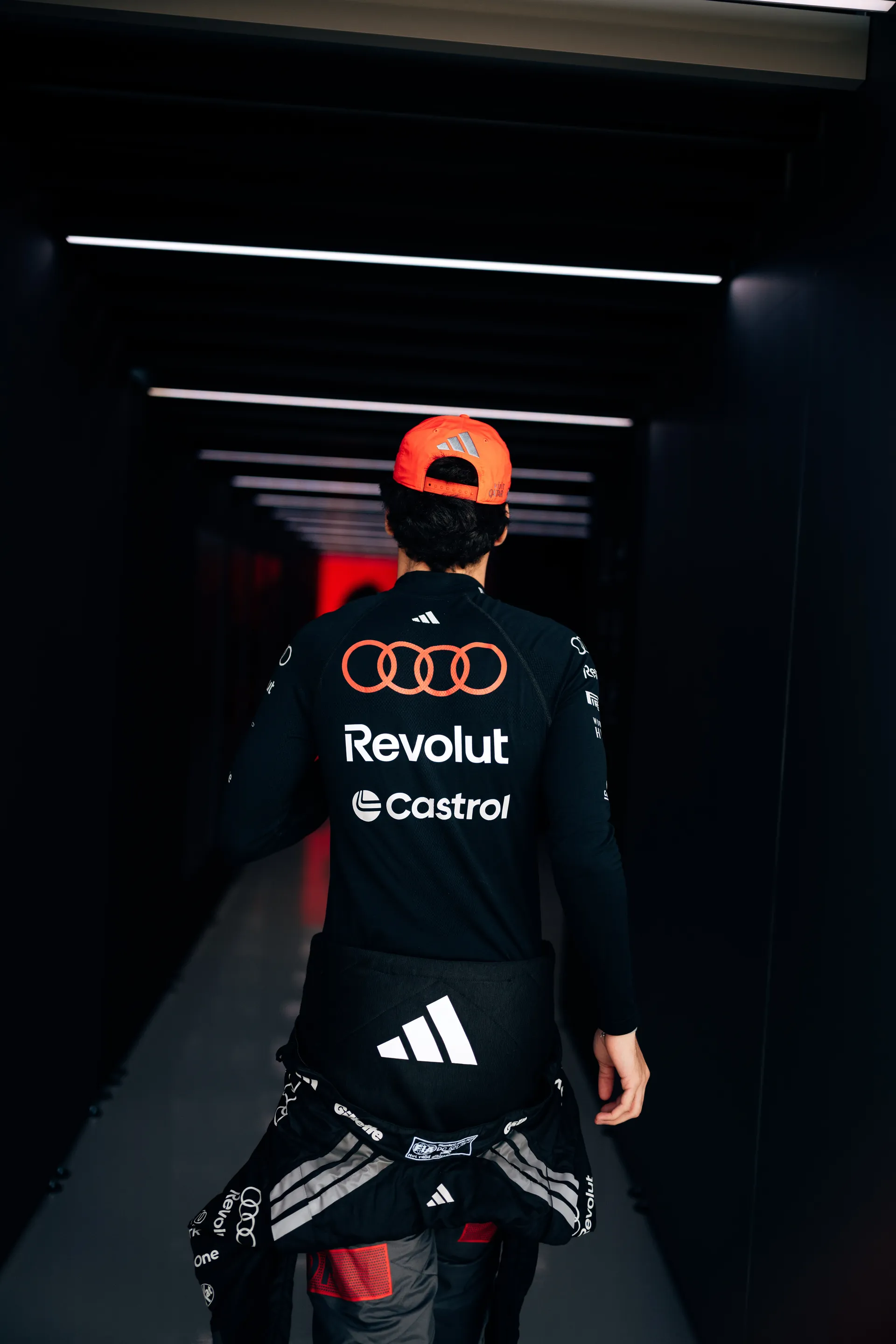 Gabriel Bortoleto walking through a dark paddock corridor in Audi Revolut F1® Team kit and an orange cap, seen from behind.