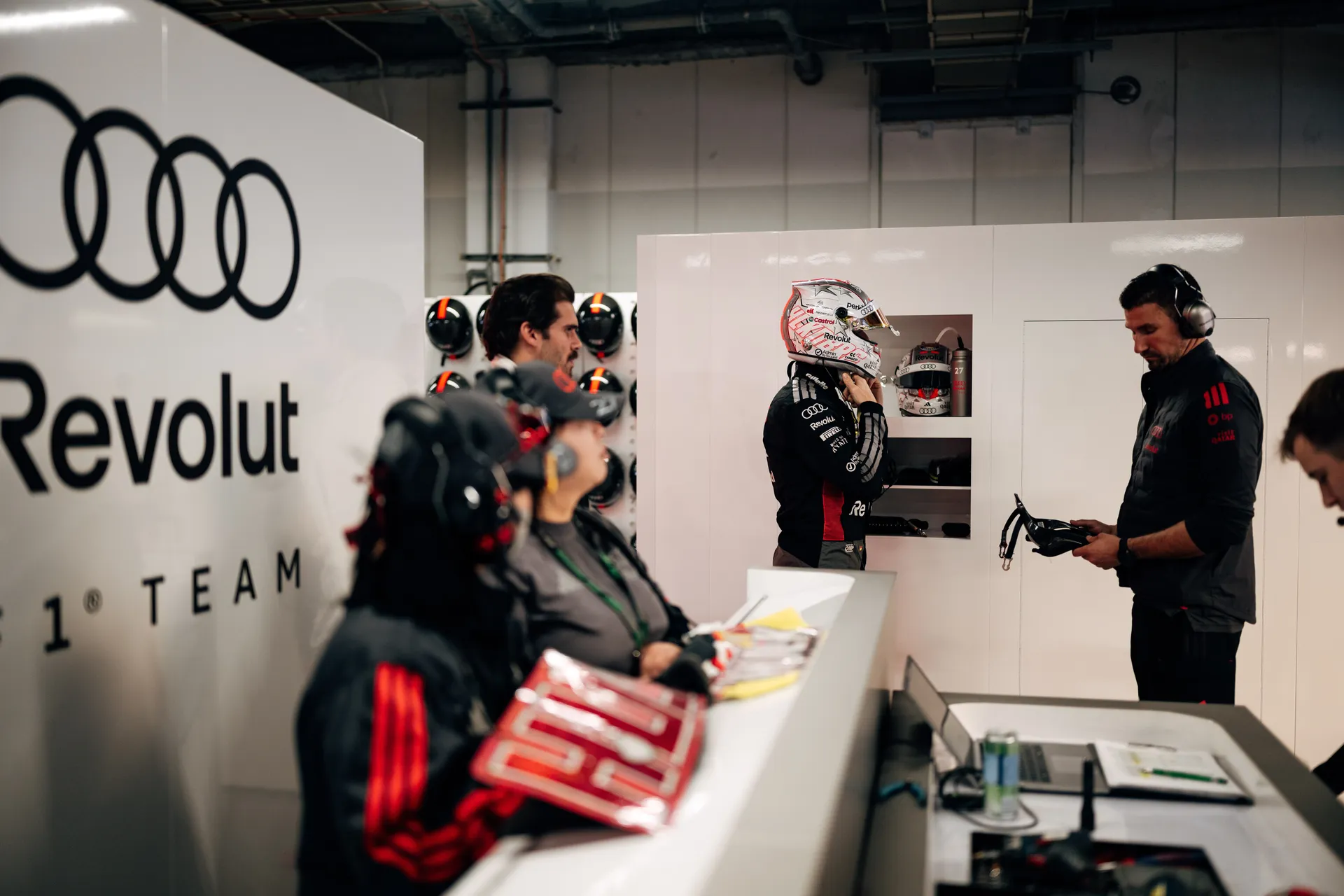 Audi Revolut F1® Team garage scene with crew on headsets while Nico Hulkenberg prepares in the background wearing his helmet.