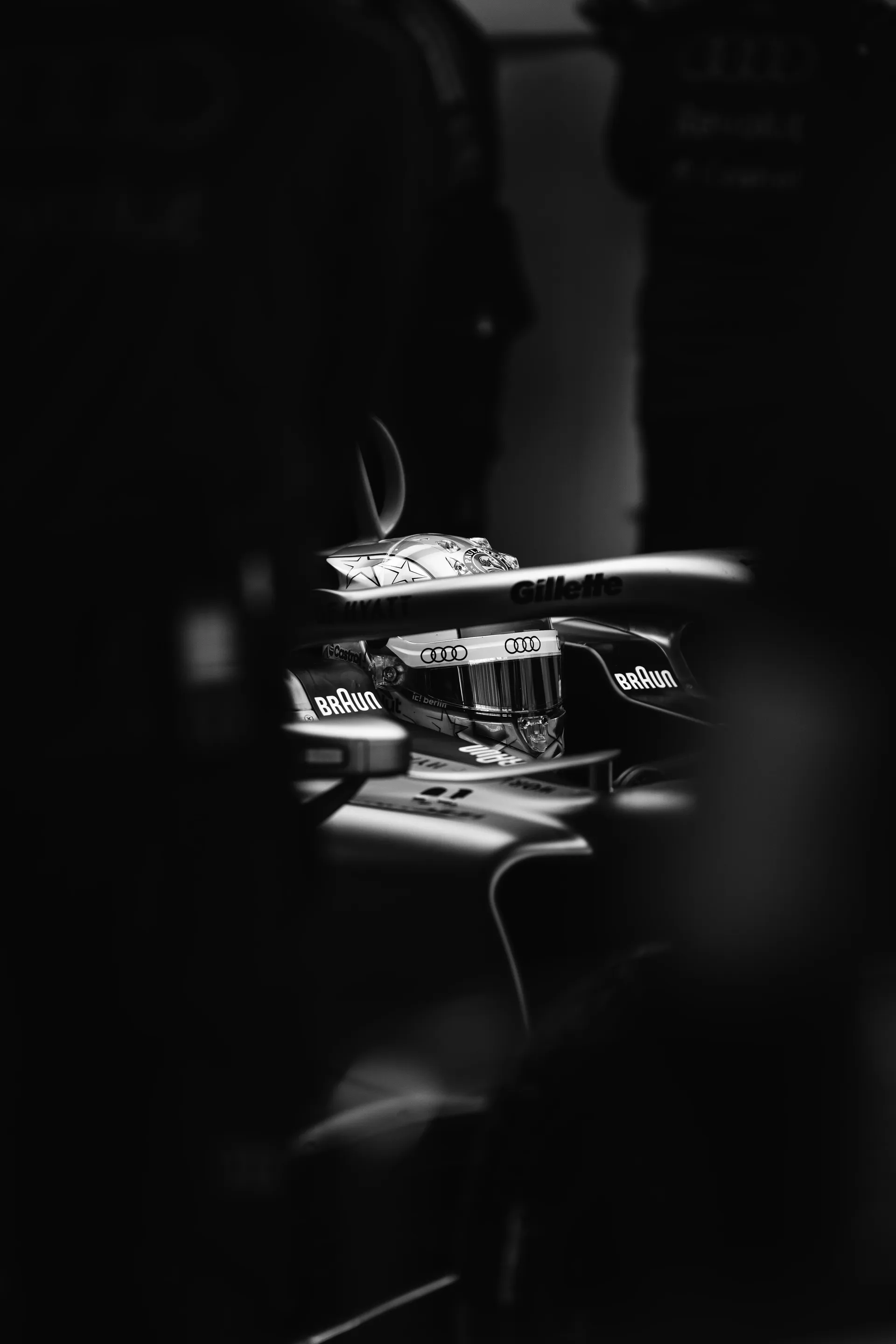 Black and white close up of Nico Hulkenberg seated in the Audi Revolut F1® Team car, seen through the halo and garage equipment, with his helmet partially framed by shadows.
